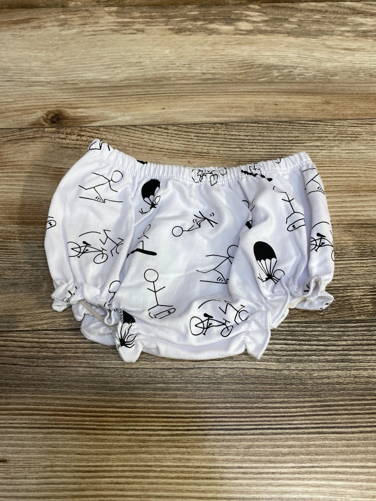 Flat lay of Stud Muffins Bloomers size 3m showcasing white fabric with black stick-figure athlete print on wood plank floor