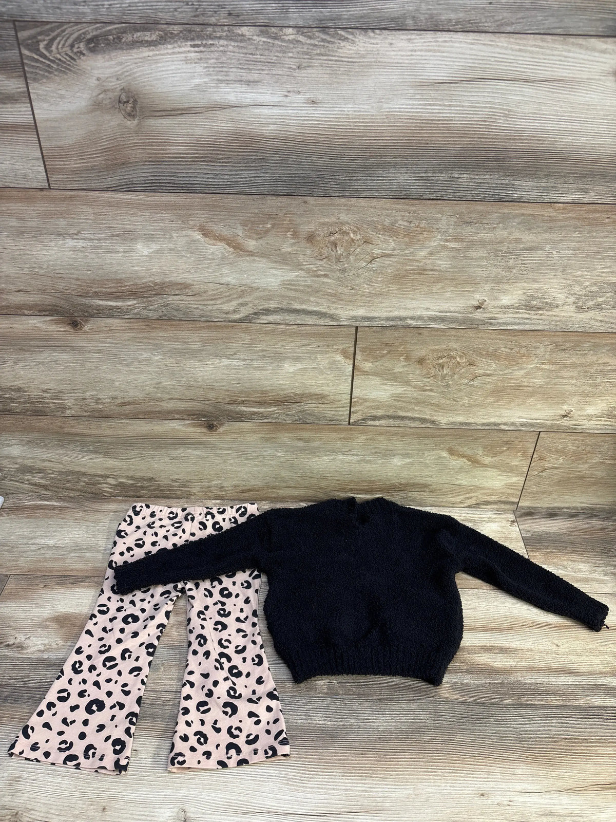 Top view of DKNY two-piece toddler set showcasing a black sweater and pink leopard-print flare pants on a wood-look floor