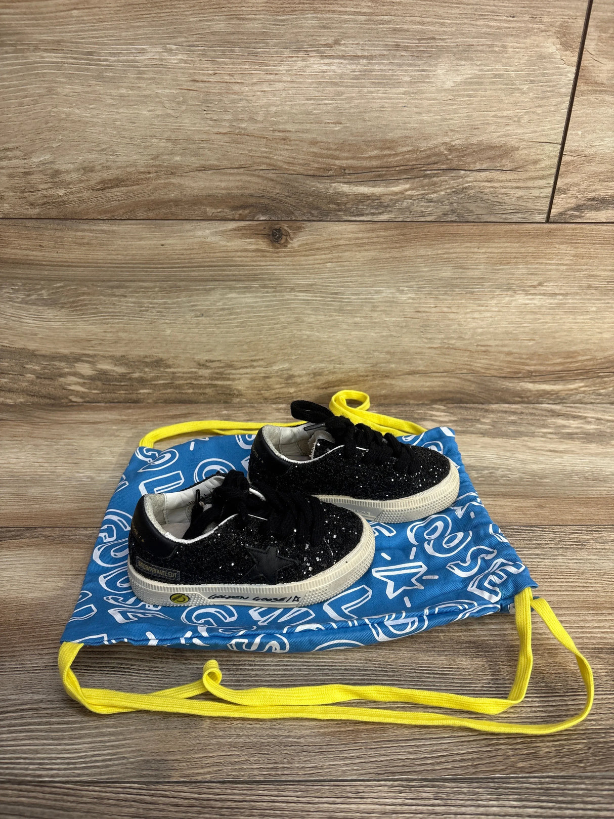 Black glittery toddler sneakers with white soles on a blue drawstring bag with white graphics and yellow straps