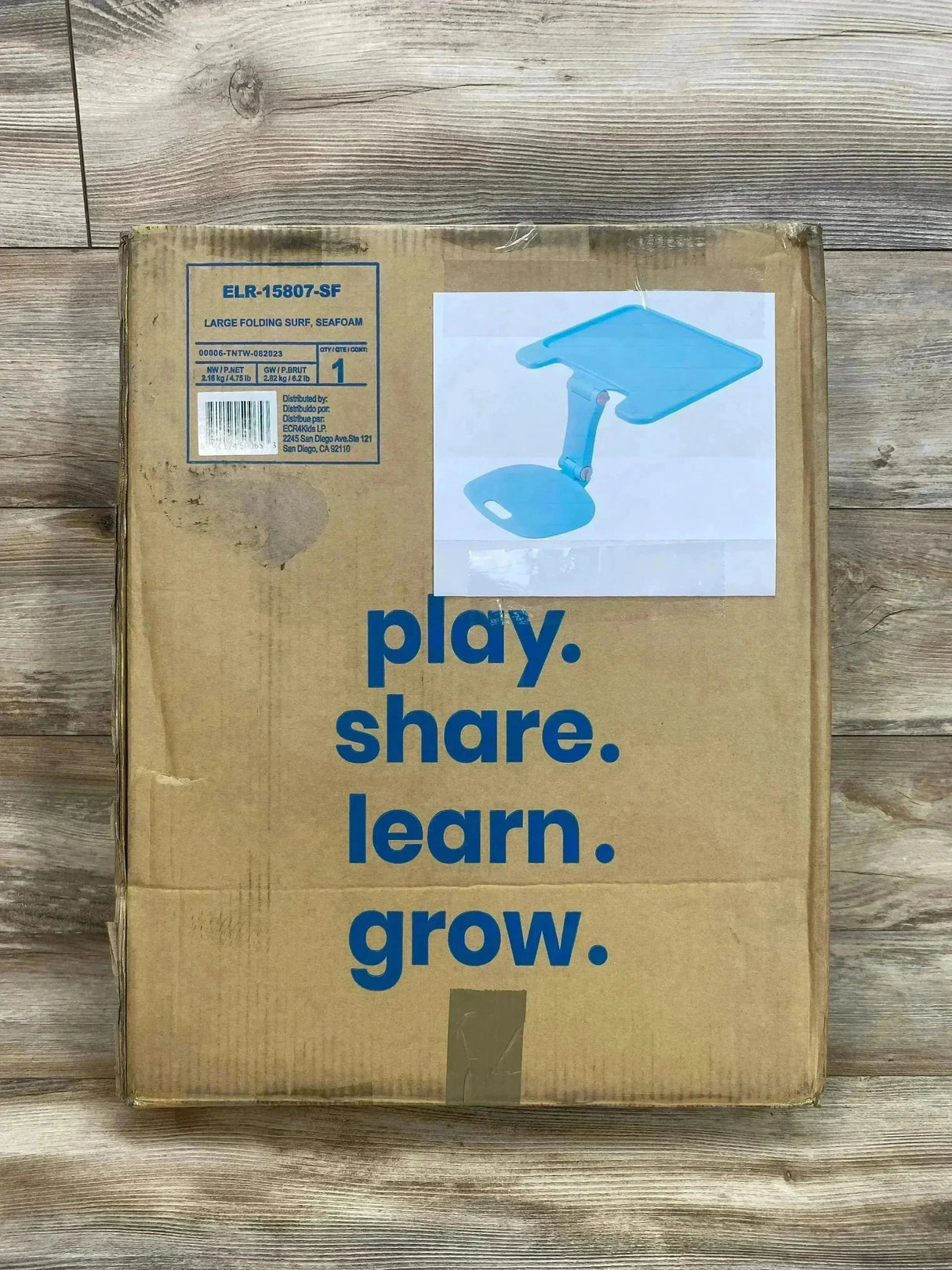 Top-down view of The Surf folding portable lap desk in seafoam, boxed showing printed text play. share. learn. grow. on the cardboard with wood floor background