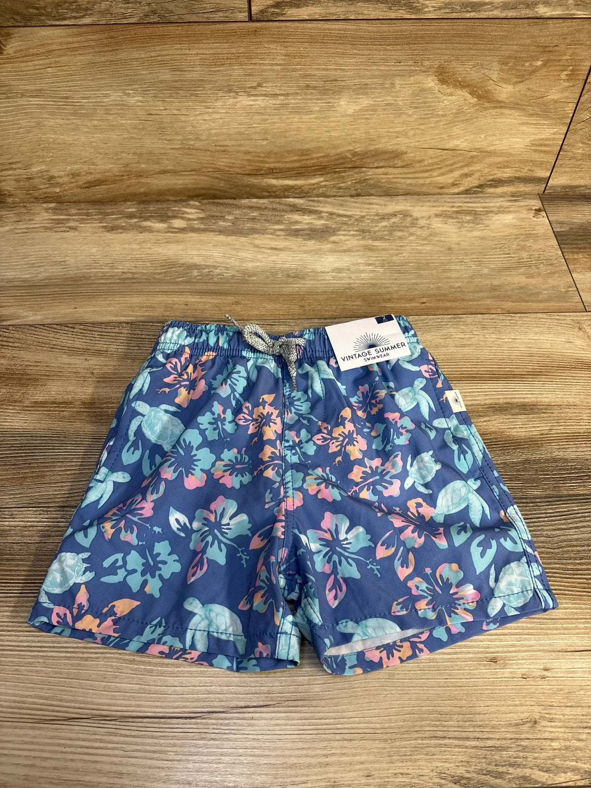 Front view of toddler blue floral swim shorts showing elastic waistband with drawstring and turtle print on wood background