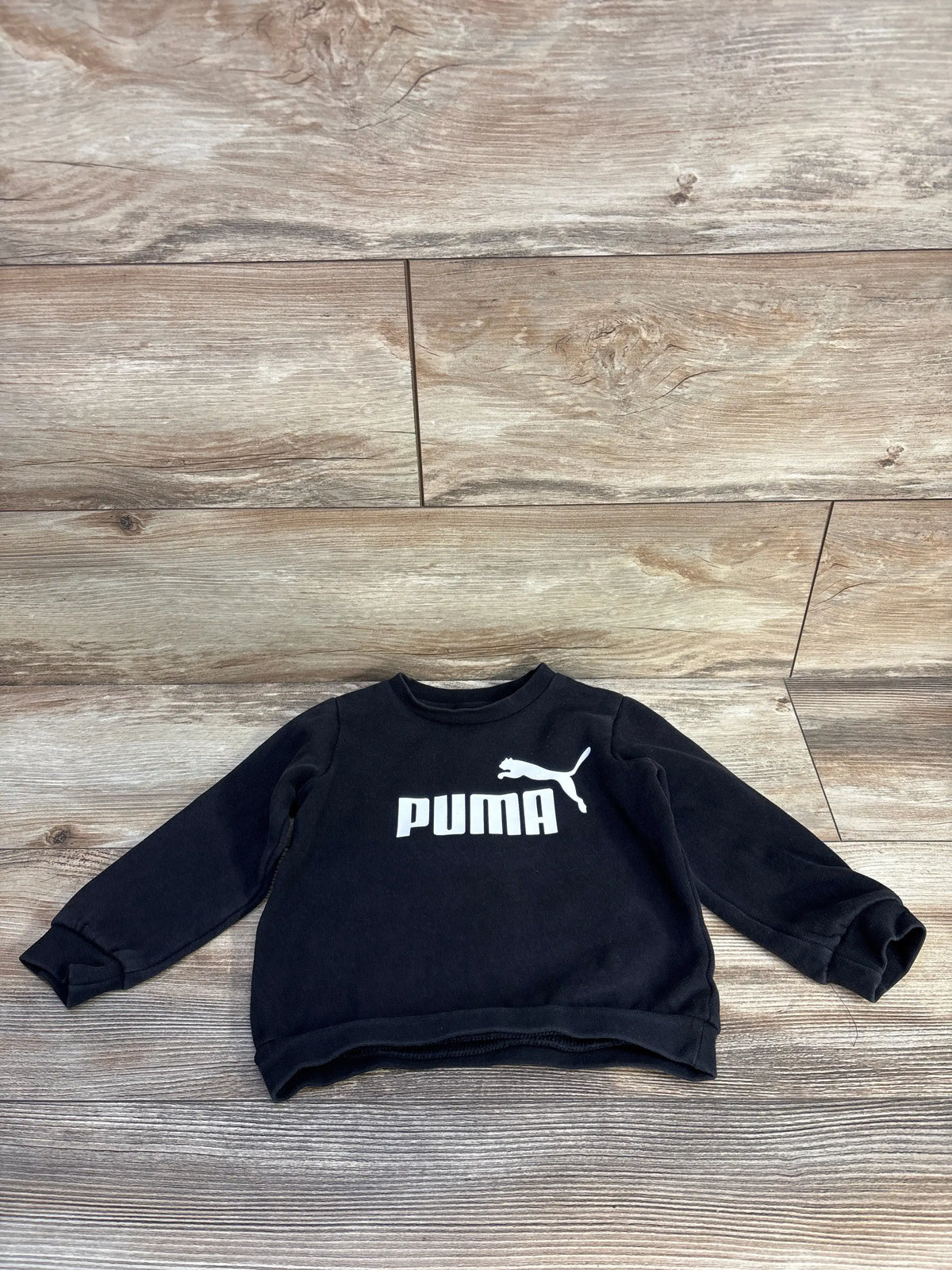 Front view of a black Puma pullover sweatshirt showcasing the white chest logo on a wood floor