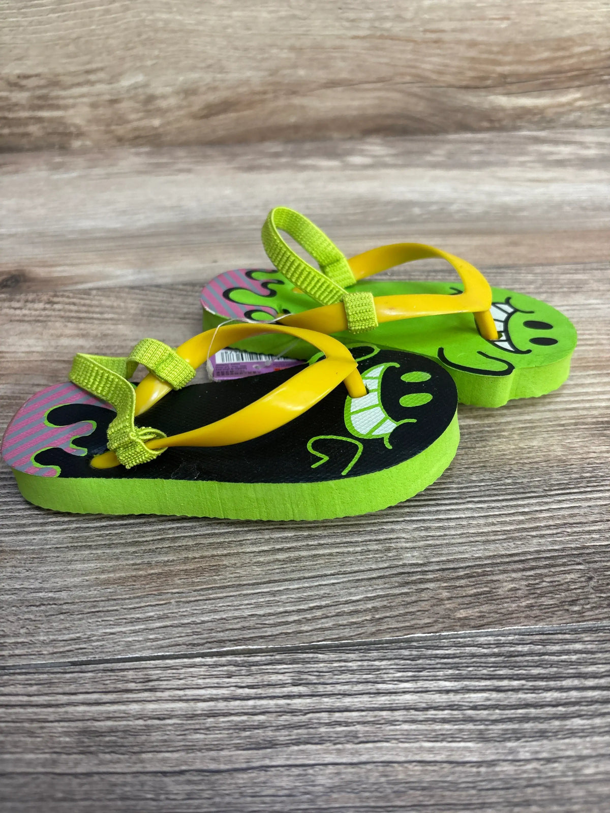 Side view of kids’ lime and black character flip-flops with yellow straps and back heel loop on wooden floor