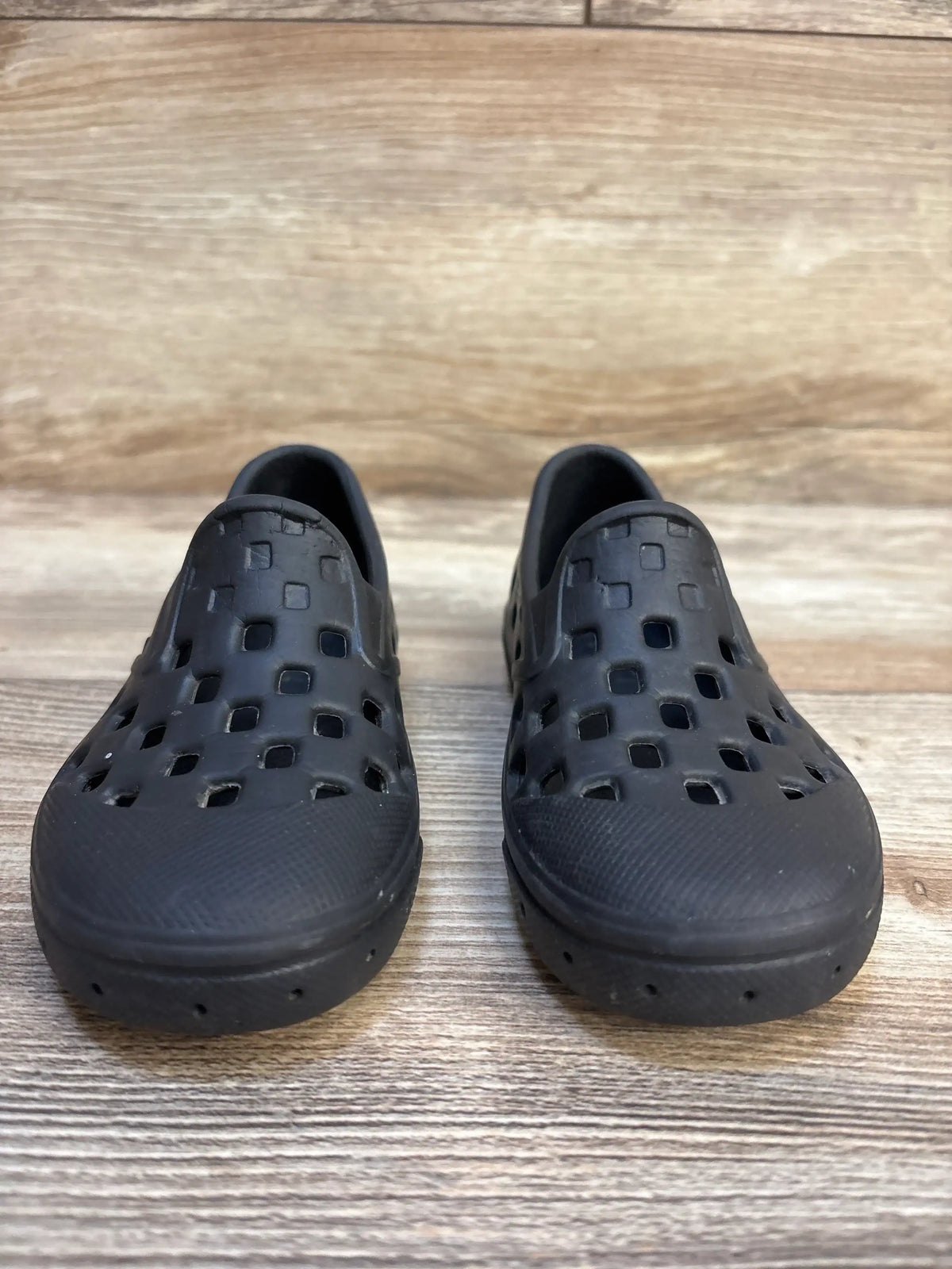 Black slip-on kids shoes with square cutouts, displayed on a wooden floor