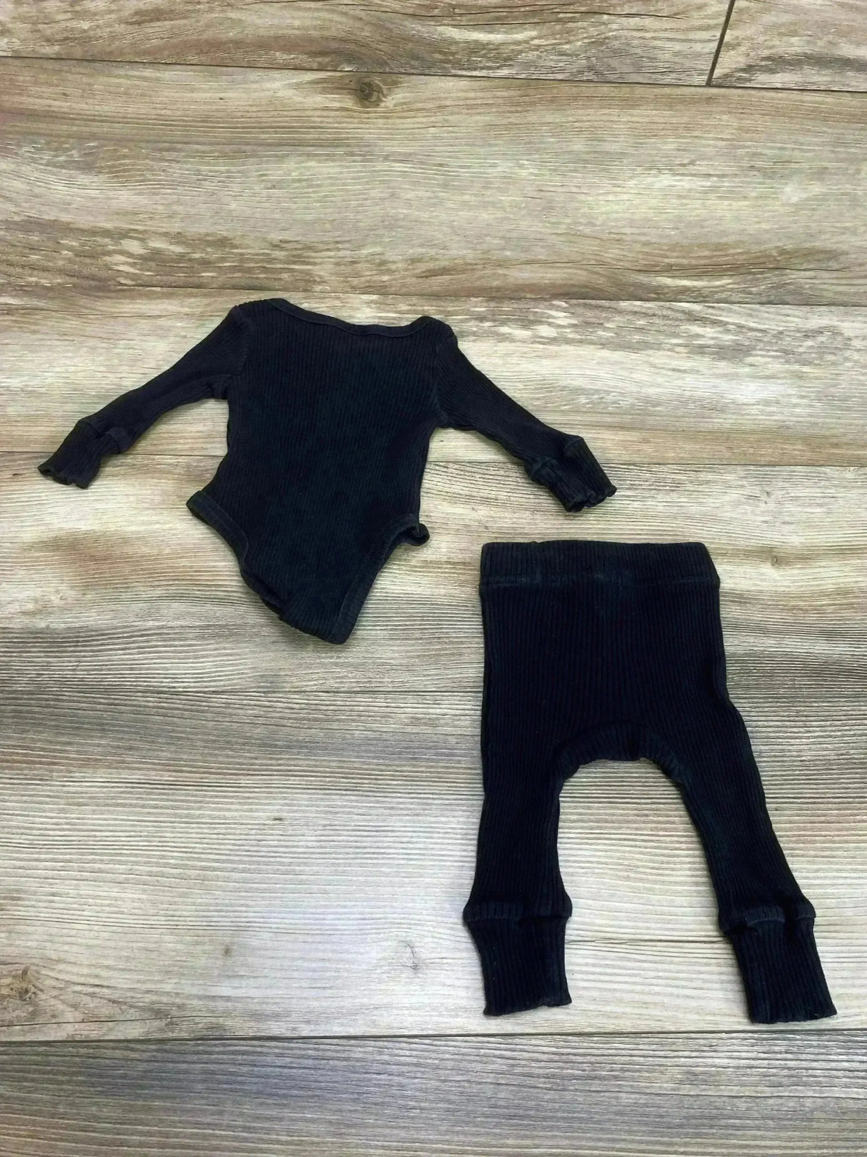 Top-down of black ribbed 2-piece baby outfit: long-sleeve bodysuit with ruffle cuffs and matching ribbed pants on wood, 3M