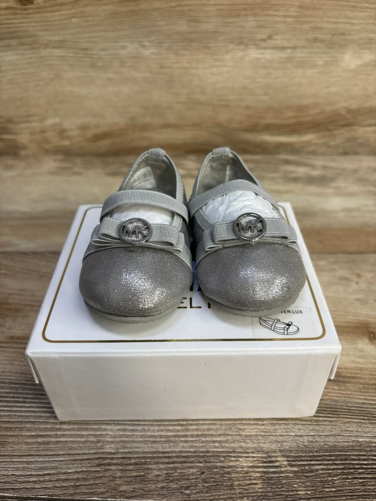 Front view of Michael Kors Rover Lux baby shoes showing silver glitter, MK logo charm and elastic strap on a white box