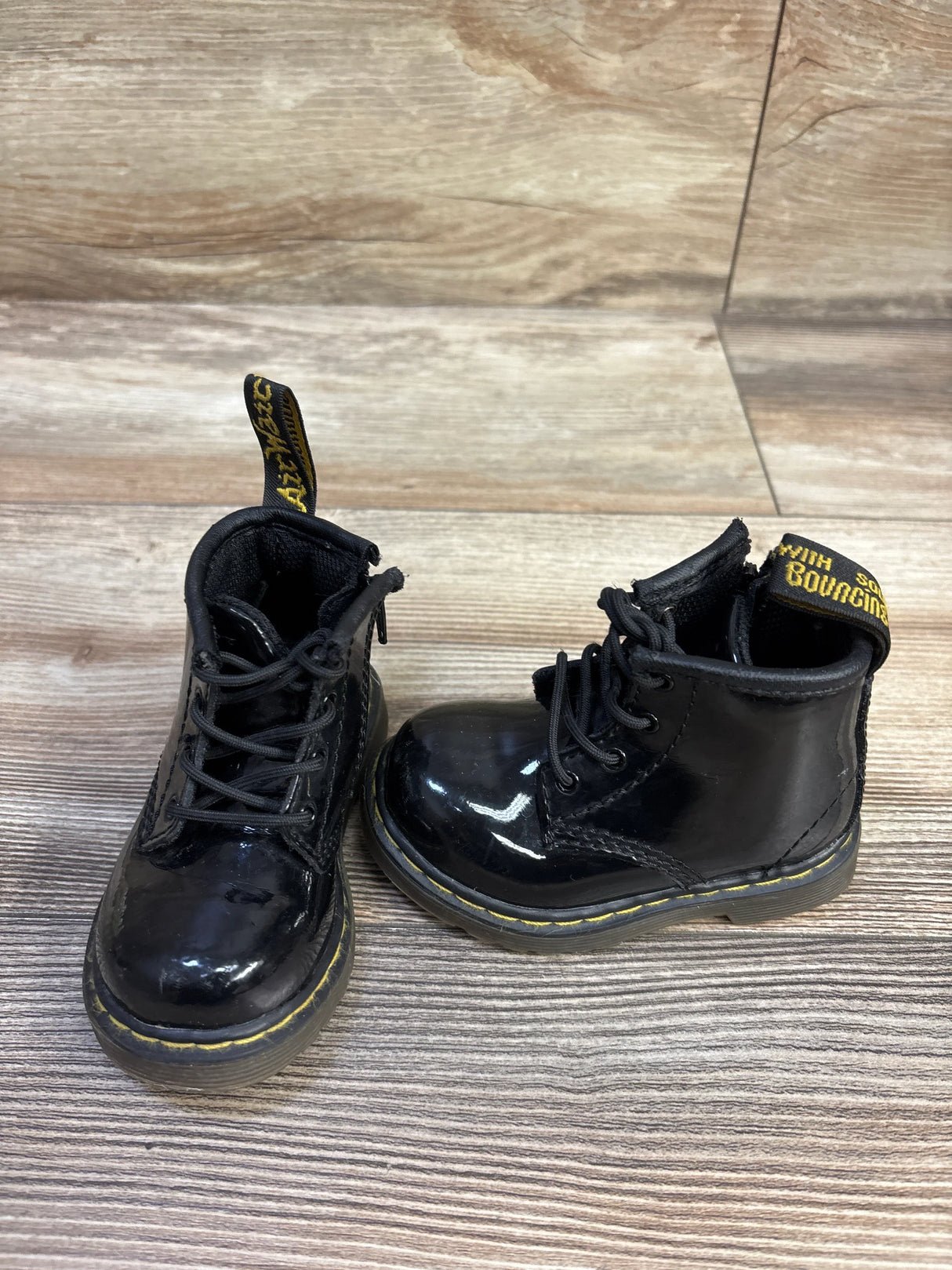Front view of toddler Dr. Martens patent leather lace-up boots showcasing glossy black finish, yellow welt stitching, and pull tabs on wood-grain background, size 4C