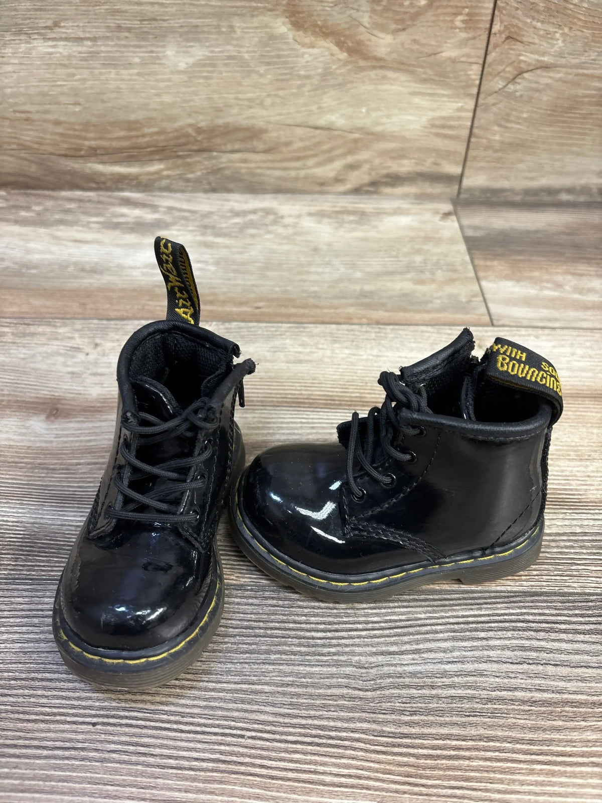 Front view of toddler Dr. Martens patent leather lace-up boots showcasing glossy black finish, yellow welt stitching, and pull tabs on wood-grain background, size 4C