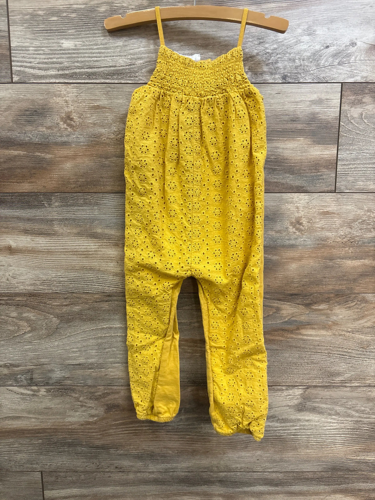 Front view of Zara yellow sleeveless eyelet jumpsuit, size 2-3T, showcasing thin straps and eyelet fabric on a wood background