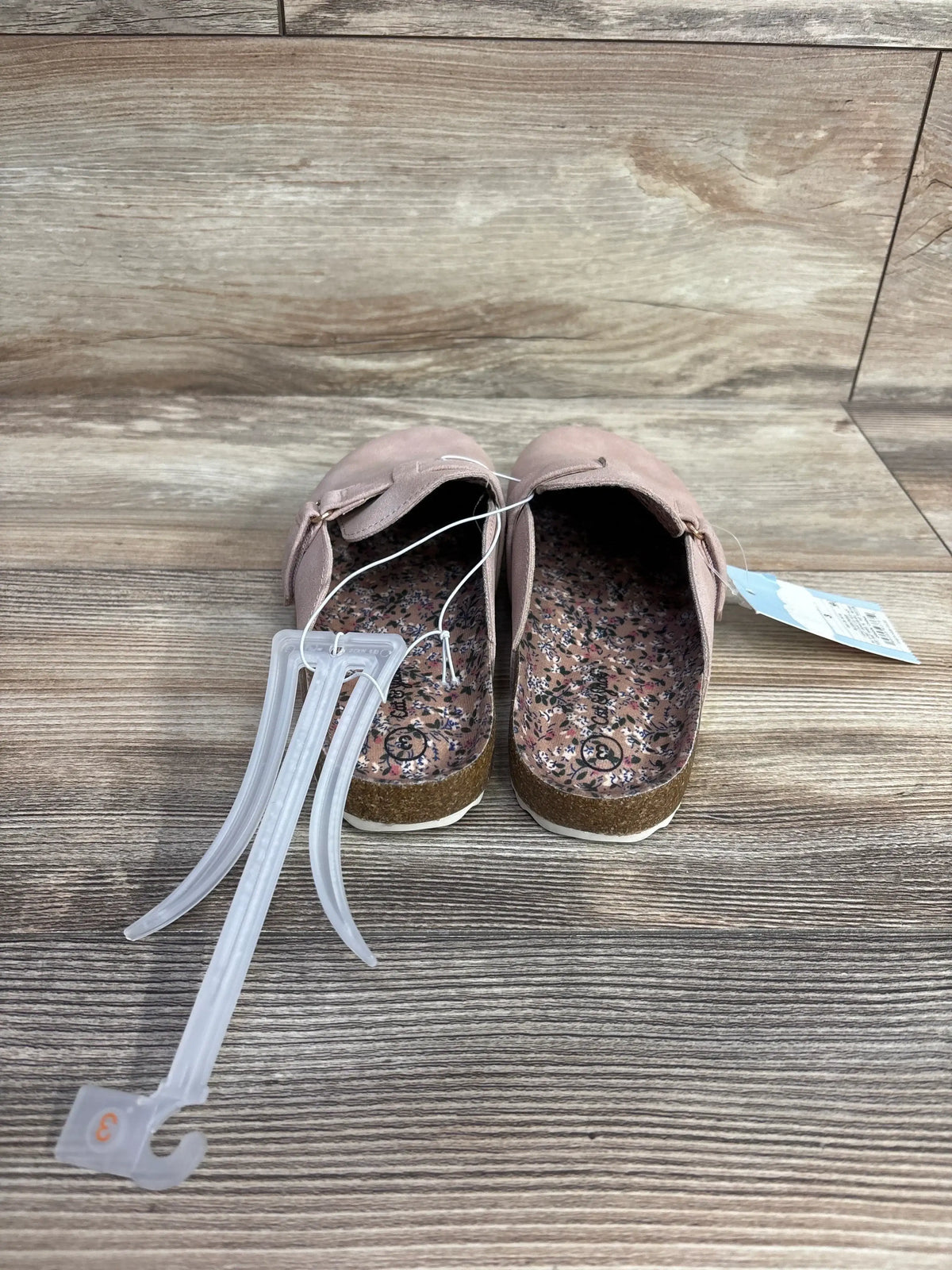 Top view of Cat & Jack Perry clogs in blush, size 3Y, showing floral insoles, adjustable strap, and attached tag on wood floor
