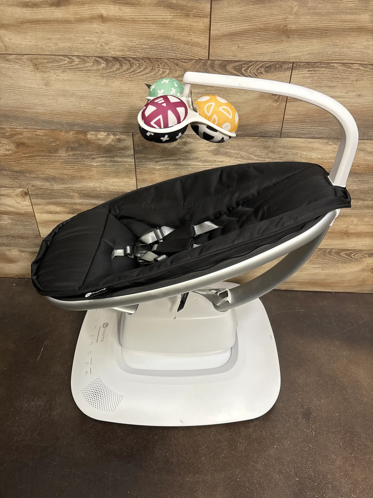 Angled view of 4moms mamaRoo baby swing showcasing black seat, harness, white base, and colorful toy bar on wood floor
