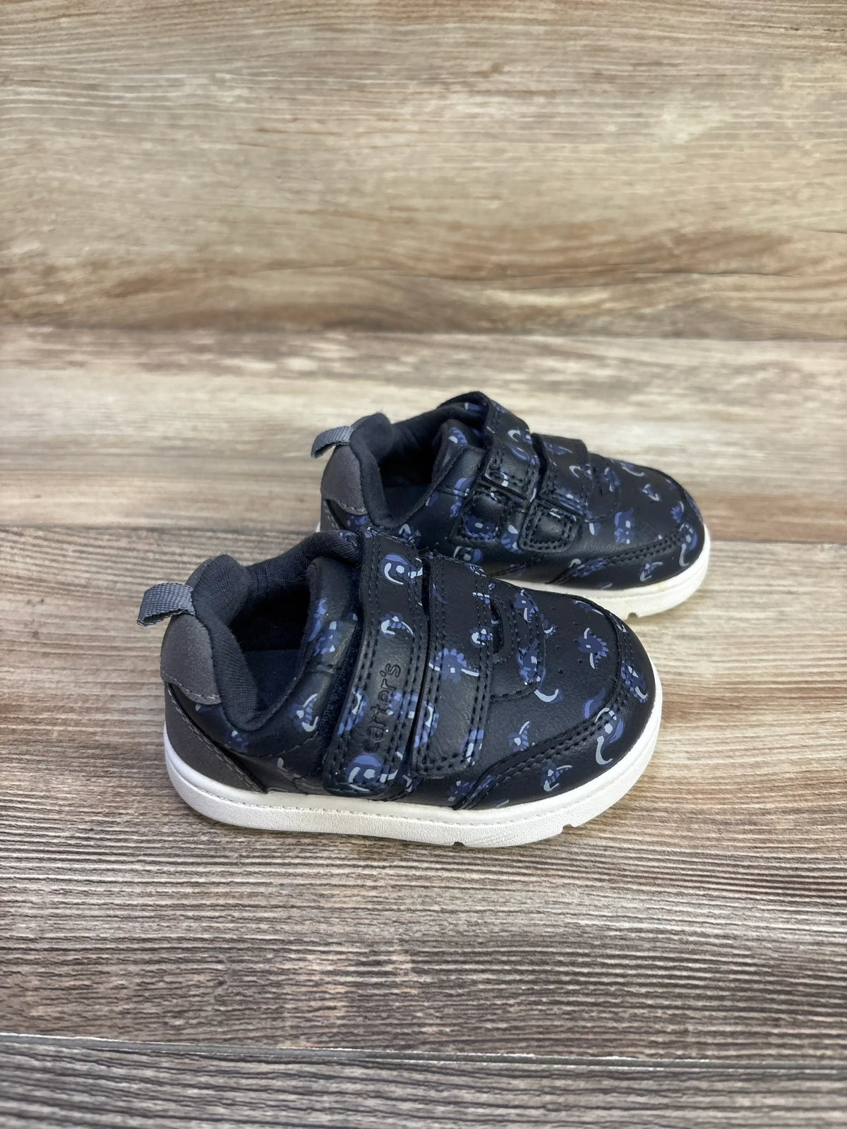 Front view of navy Carter’s Neo Dino First Walker sneakers on wood floor, showing two hook and loop straps and pull tabs