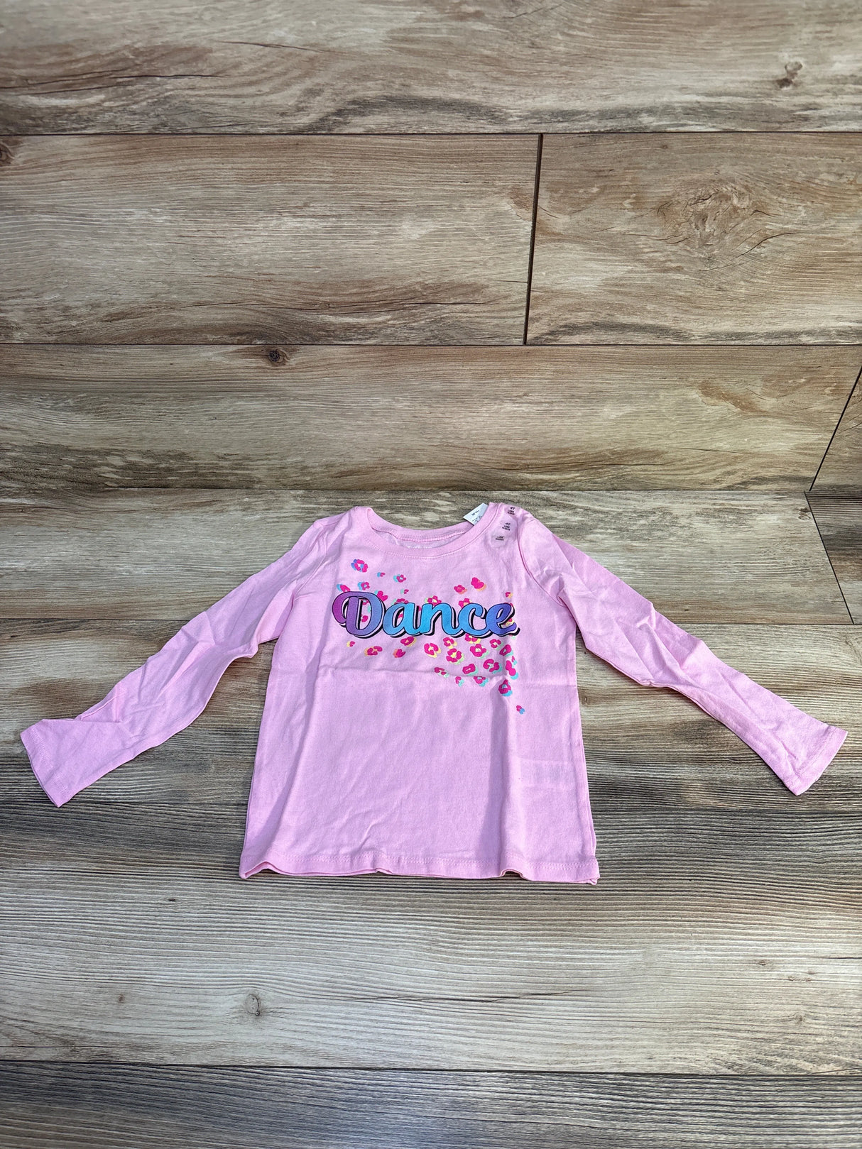 Pink long-sleeve kids shirt with "Dance" text and colorful floral print on wooden floor