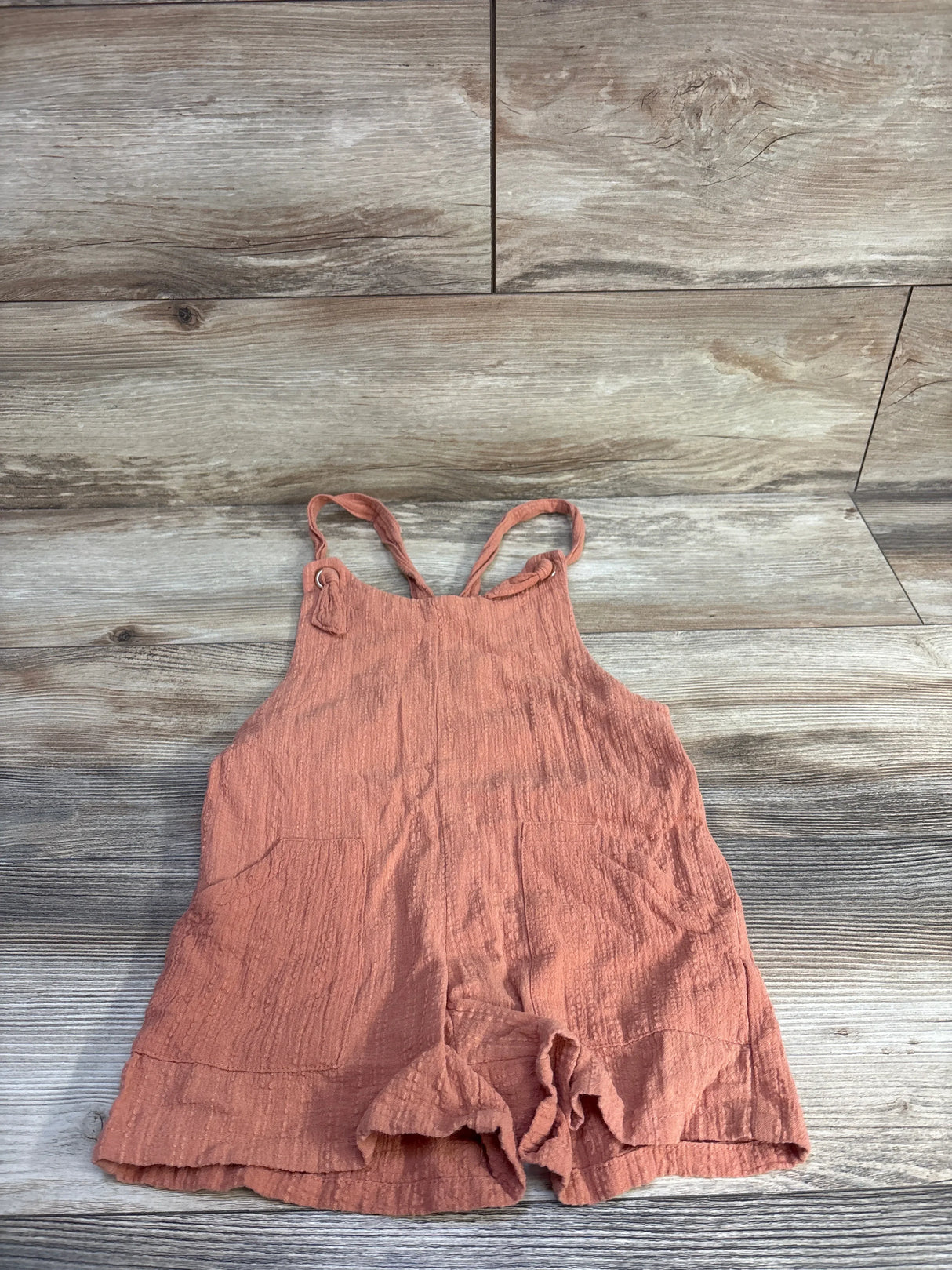 Front view of coral gauze shortalls, size 4/5, crinkled fabric, tie shoulder straps, pockets, ruffled hem on light wood