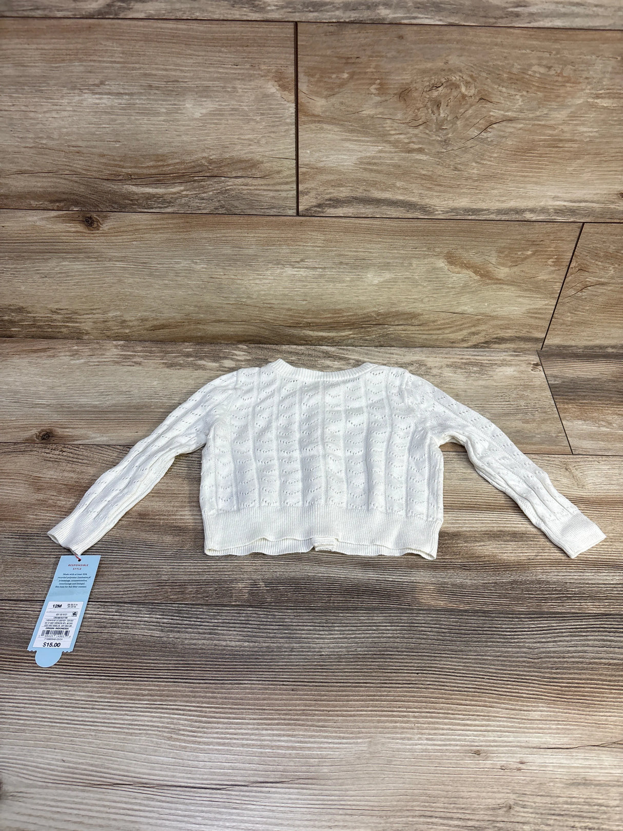 Flat lay of cream Cat and Jack pointelle baby cardigan size 12M showing knit pattern, sleeves, and attached tag on wood floor