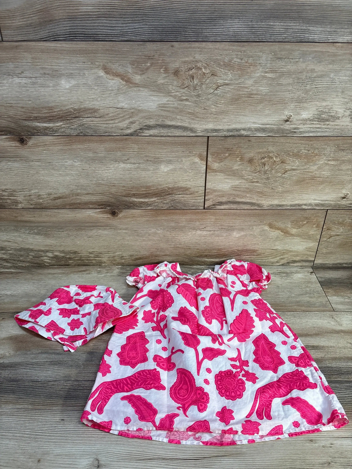 Flat lay of white toddler dress and matching hat showing bright pink floral print and puff sleeves on wood background