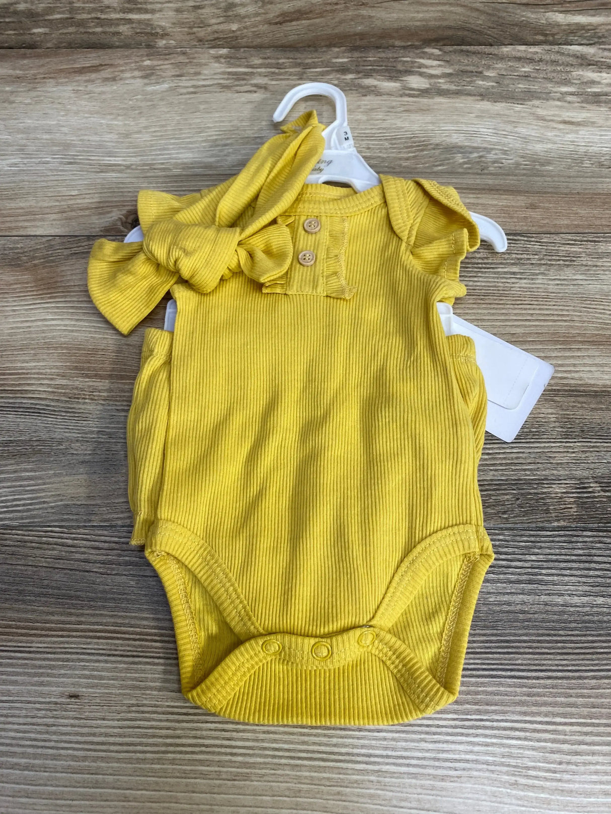 Front view of yellow ribbed baby bodysuit showcasing ruffle sleeves and snap crotch with matching knotted headband on wood background