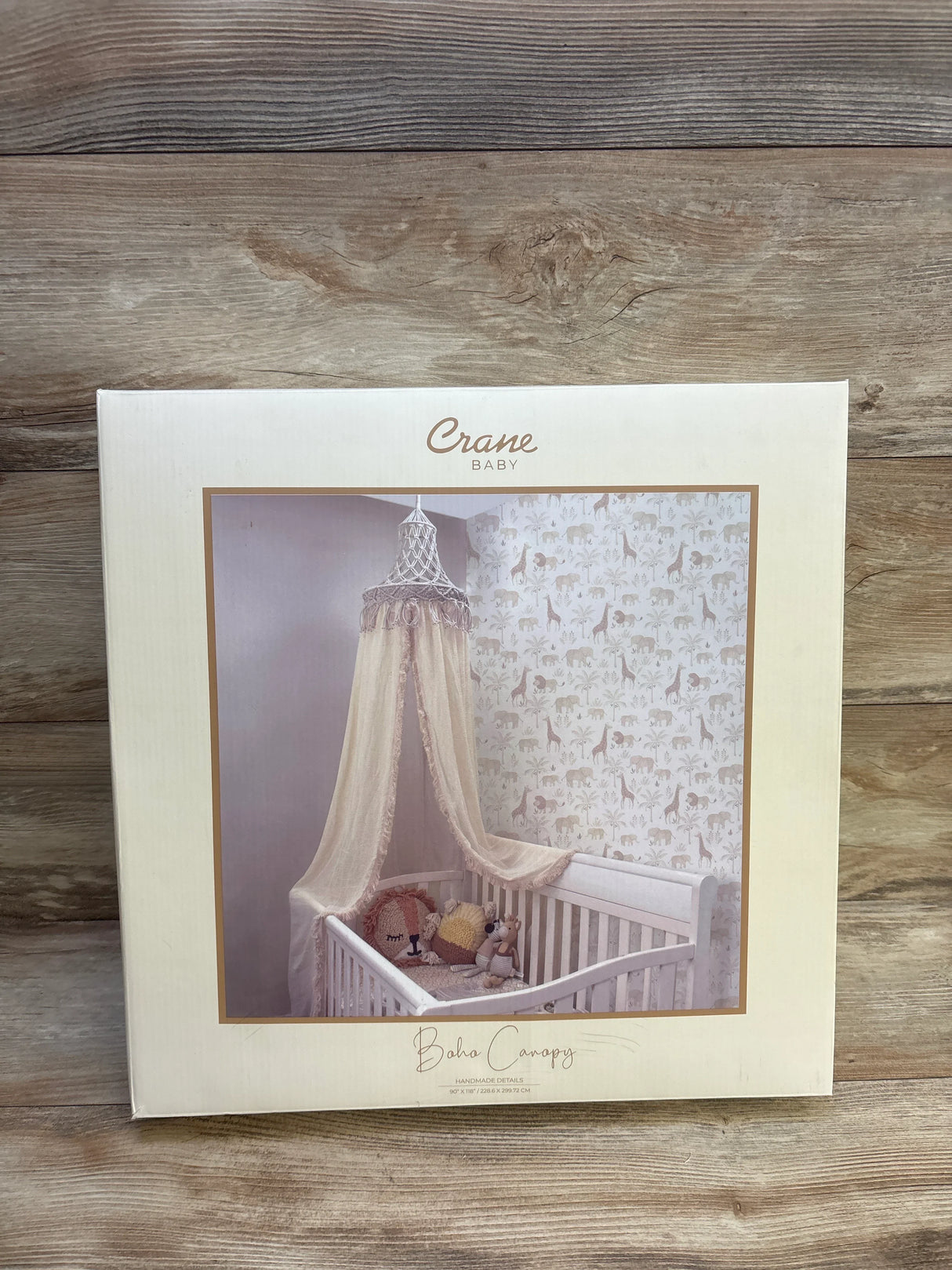 Front view of Crane Baby Boho crib canopy box showcasing macrame top and sheer draped canopy over a white crib with wood plank background