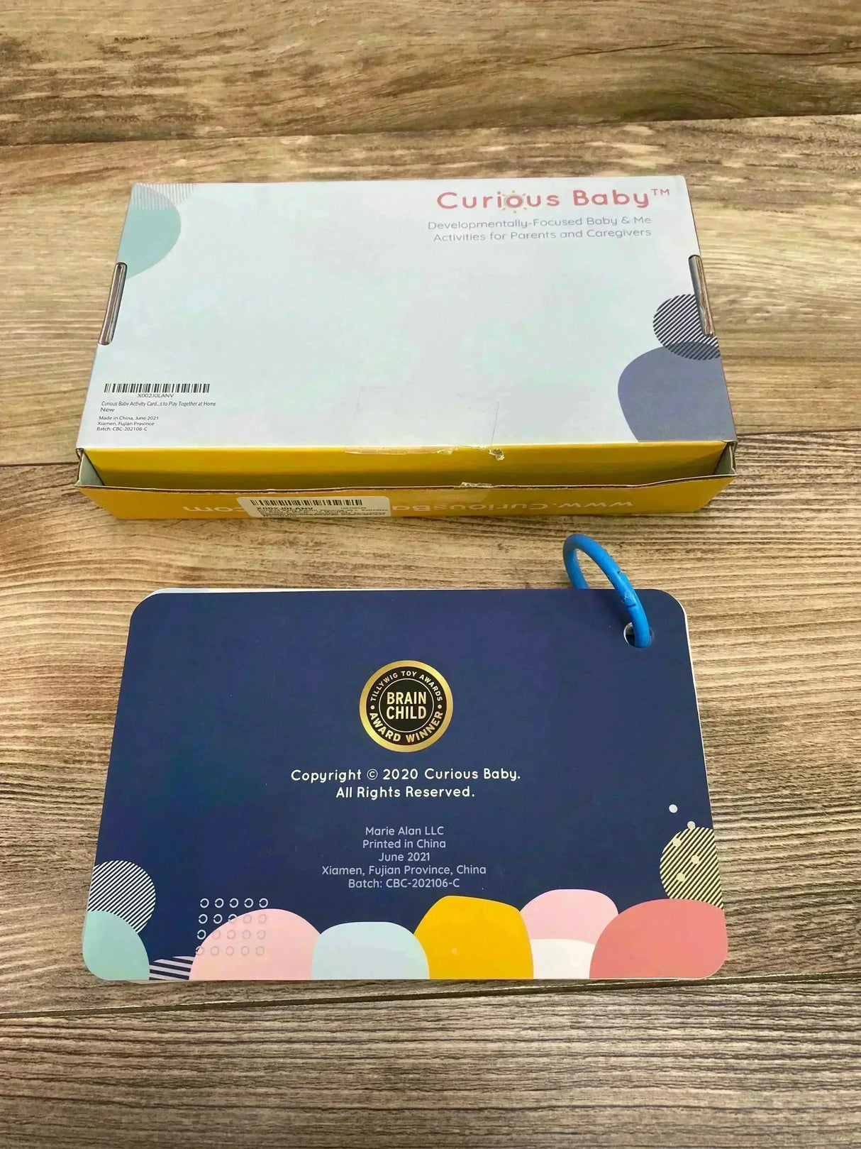 Overhead view of Curious Baby cards with box showing navy card with blue ring and gold Brain Child award seal on wood table