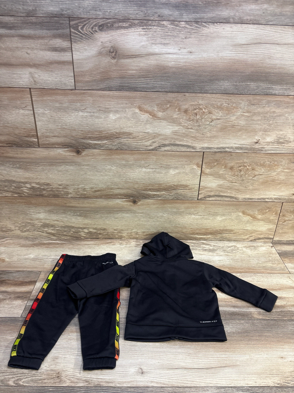 Flat lay of Nike 2-piece tricot Therma-Fit hooded tracksuit set (size 12M) showing black hoodie and jogger pants with multicolor side stripes on wood background