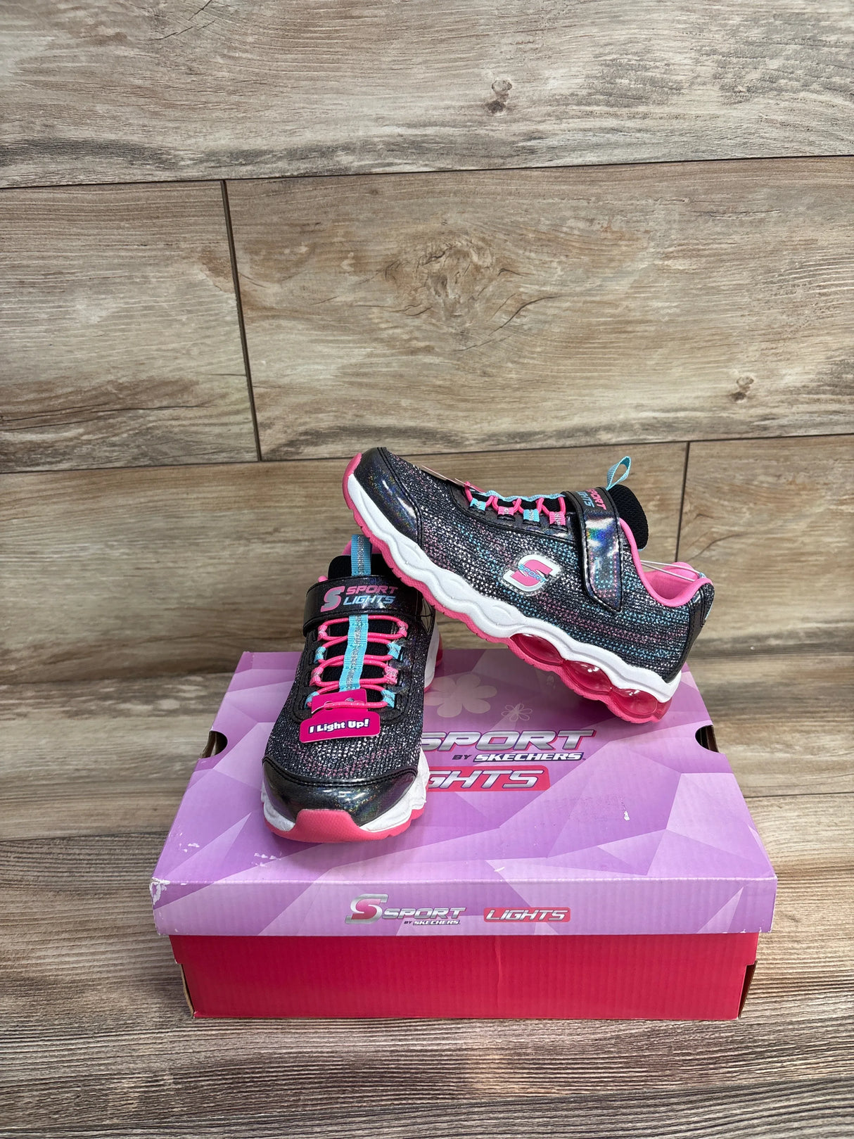 Black and pink Skechers Sport Lights kids shoes with light-up feature on pink retail box against wood background