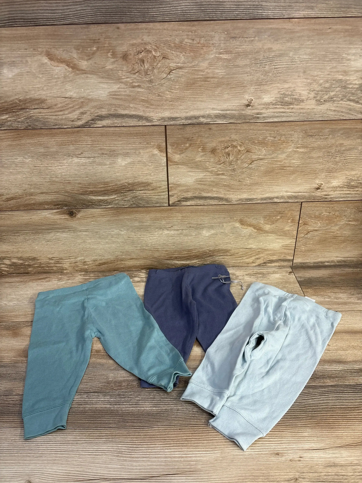 Flat lay of Cloud Island 3-pack pull-on baby pants in mint, navy, and pale blue with cuffed ankles on wood background