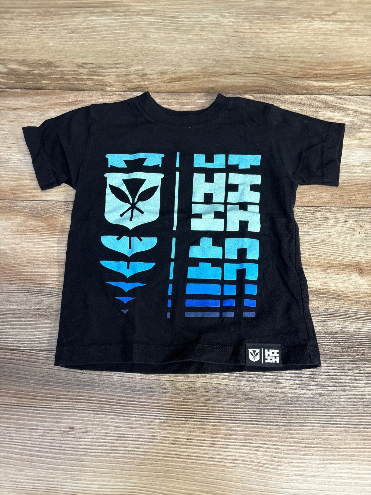 Black kids t-shirt with blue geometric graphics and leaf emblem, on wood background