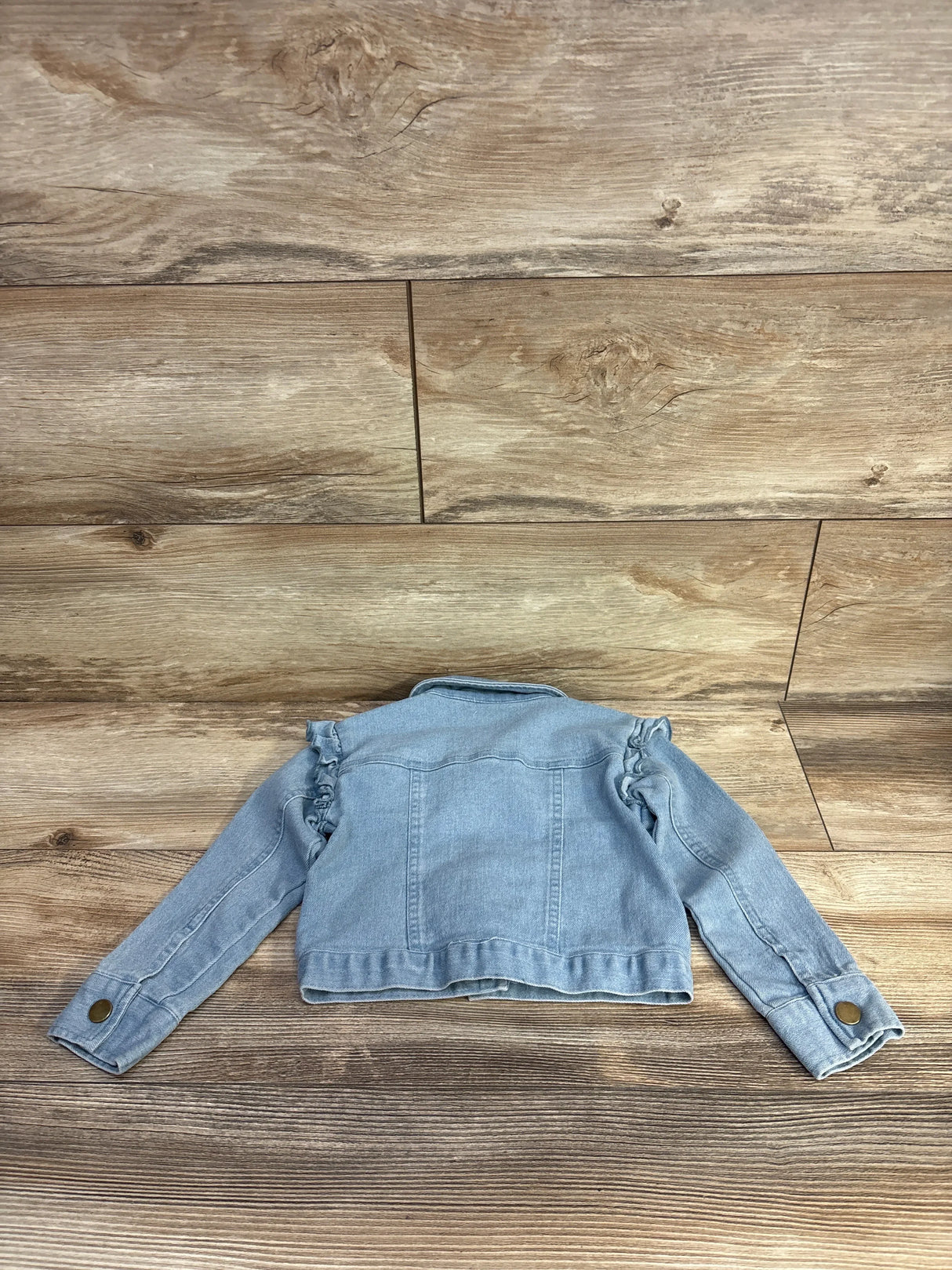 Back view of Btween light blue denim jacket showcasing ruffled shoulders and buttoned cuffs on wood background