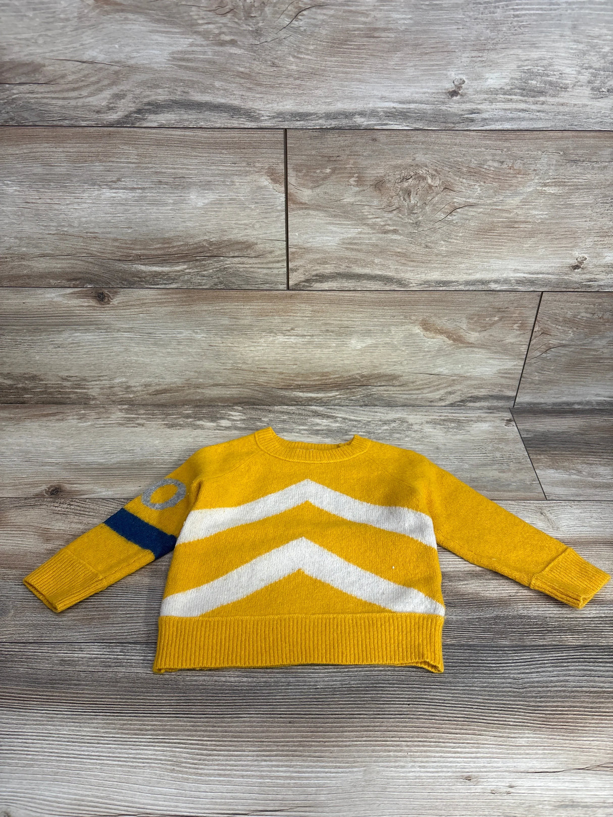 Yellow kids sweater with white chevron stripes and blue circle, laid on wooden floor