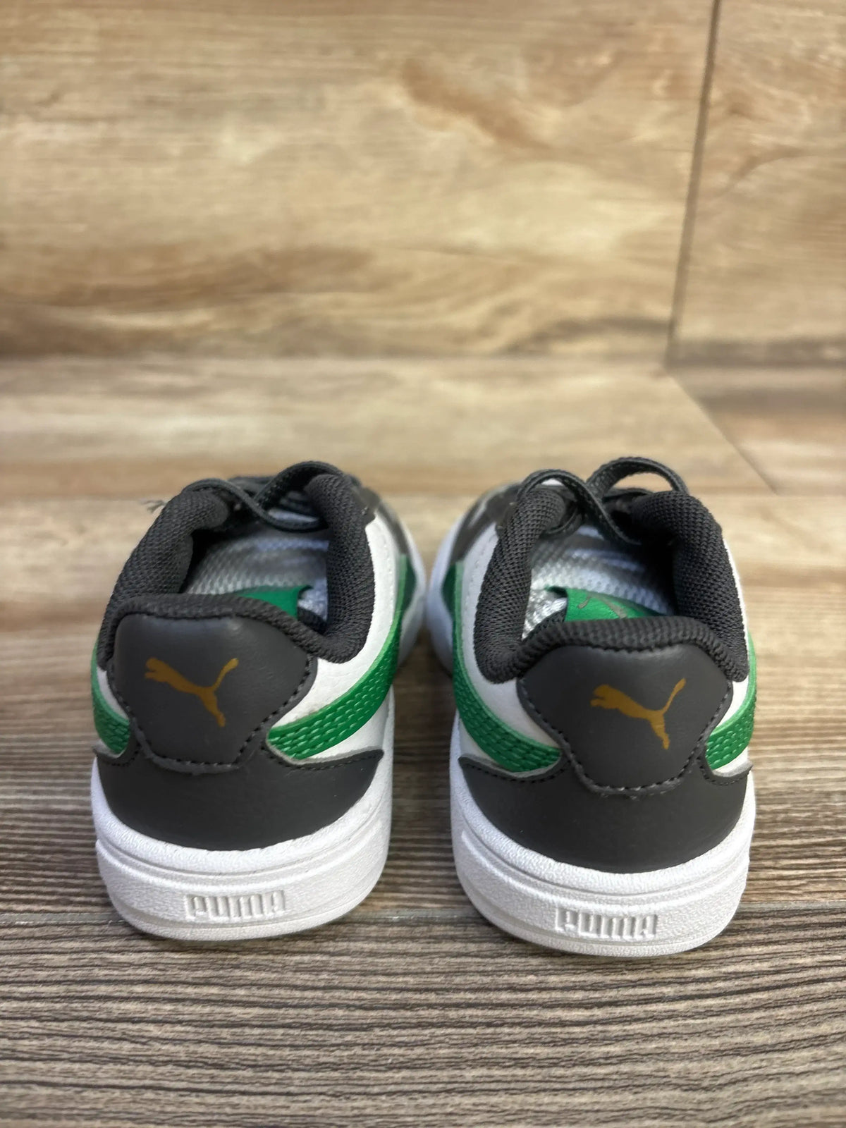 Rear view of Puma Caven 2.0 sneakers showing black heel with gold Puma logo, green stripe and white sole on wood floor