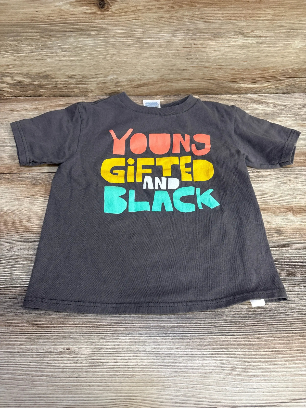 Front flat-lay of grey toddler T-shirt (4T) showcasing large colorful "Young Gifted and Black" graphic on a light wood background