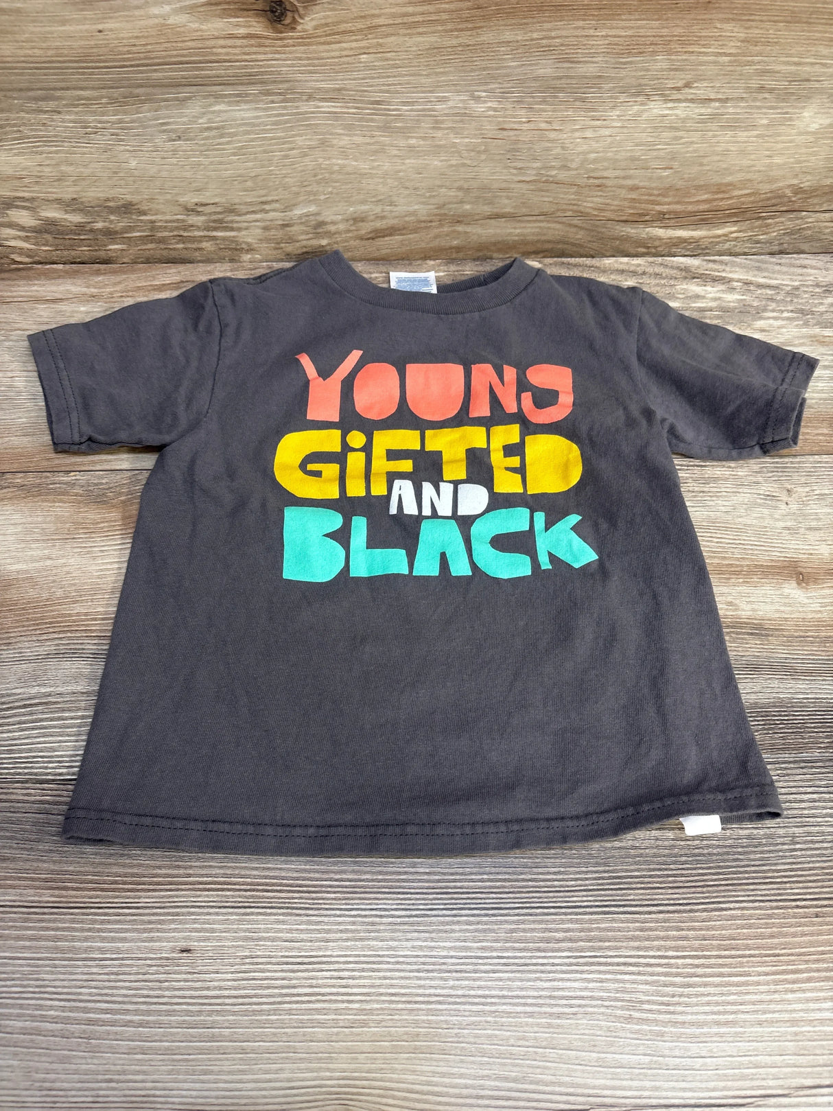 Front flat-lay of grey toddler T-shirt (4T) showcasing large colorful "Young Gifted and Black" graphic on a light wood background