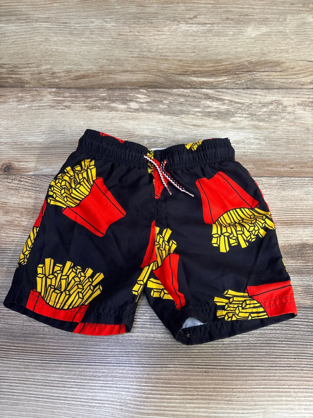 Flat lay of boys' Appaman swim trunks with red and yellow french fry print on black, elastic waistband, wood floor background