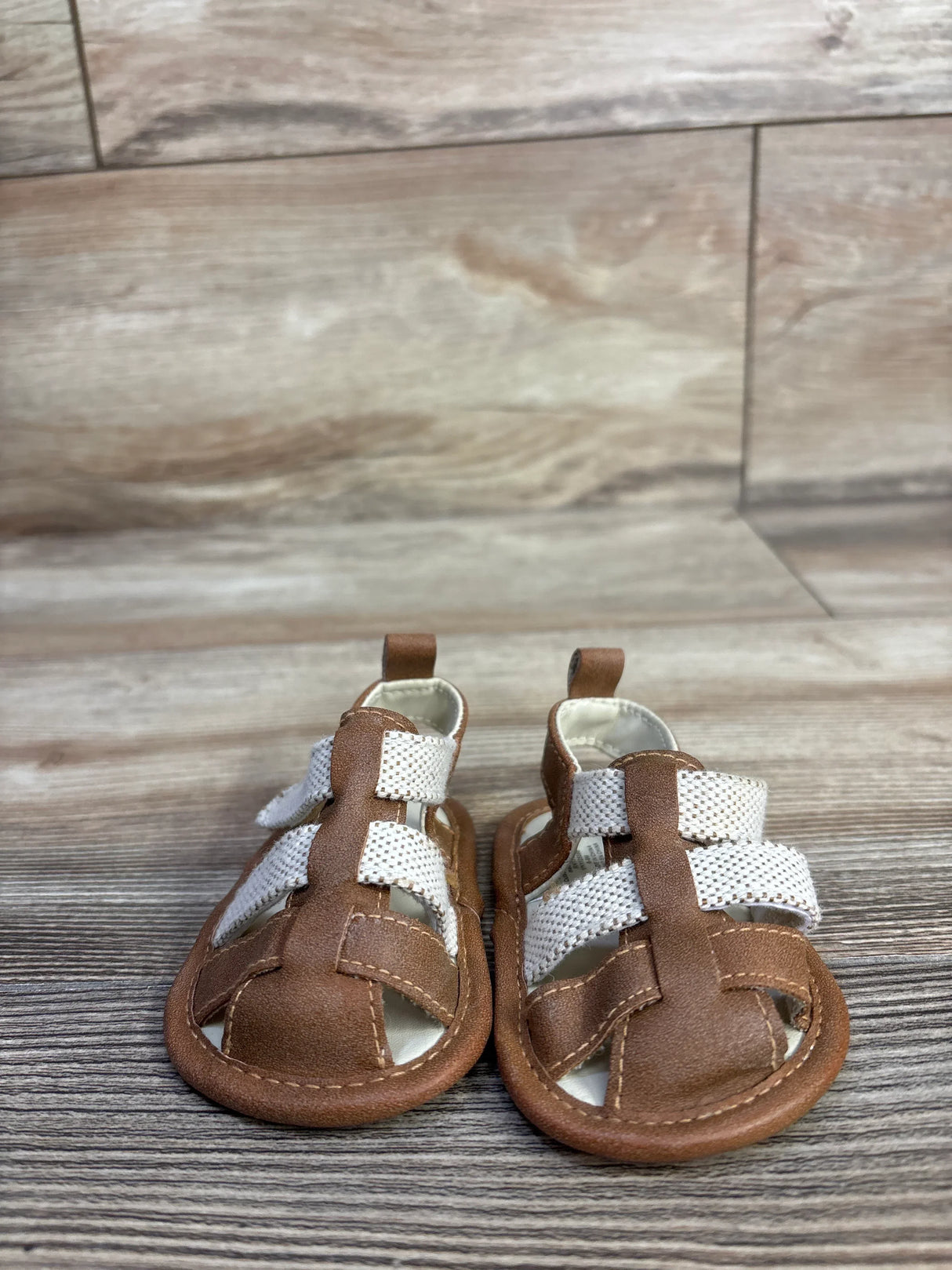 Front view of Cat and Jack tan soft-sole baby sandals showcasing white woven straps and brown faux-leather upper on woodgrain background