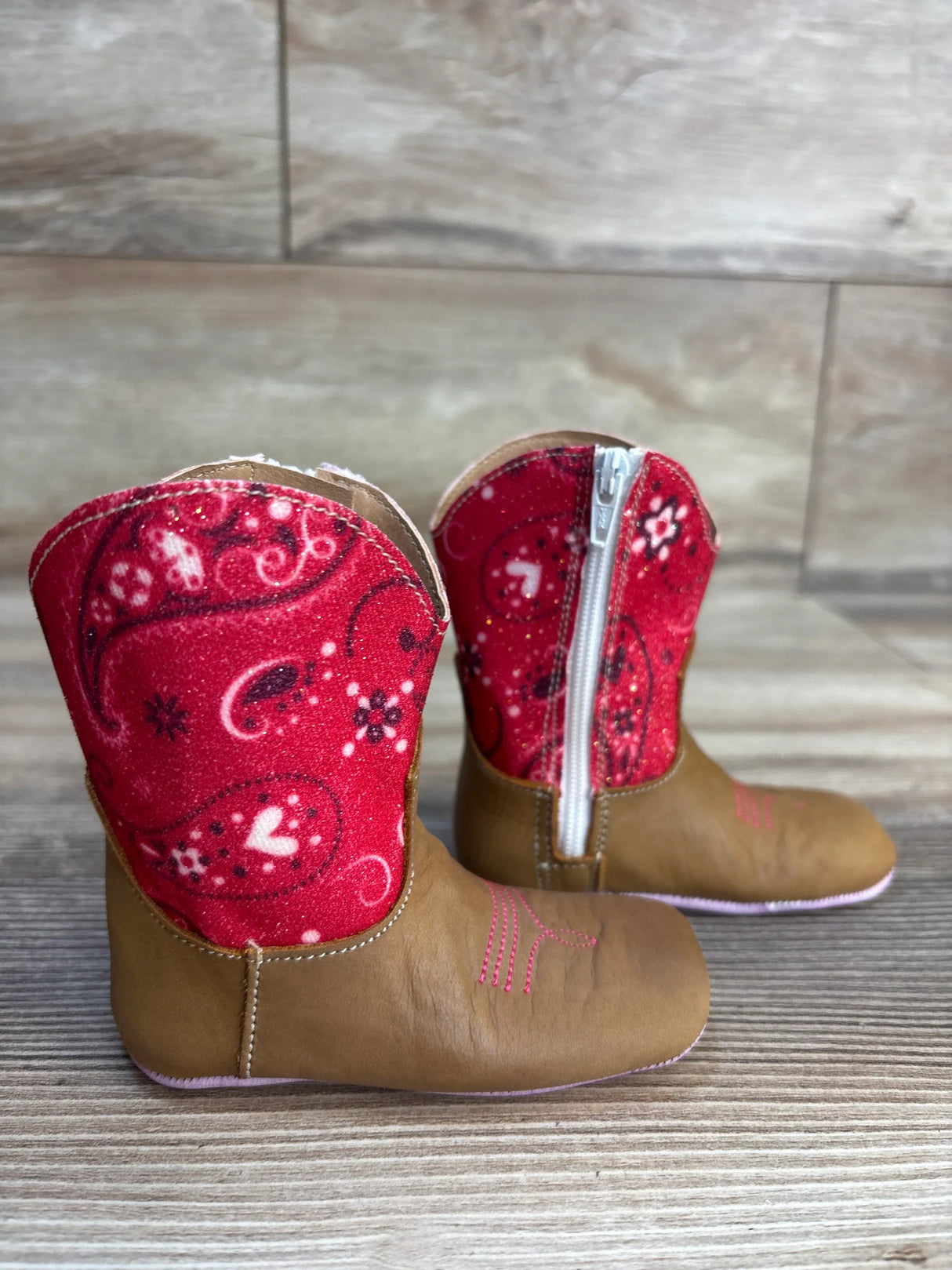 Infant brown cowboy boots with red paisley fabric and zipper on wood background.