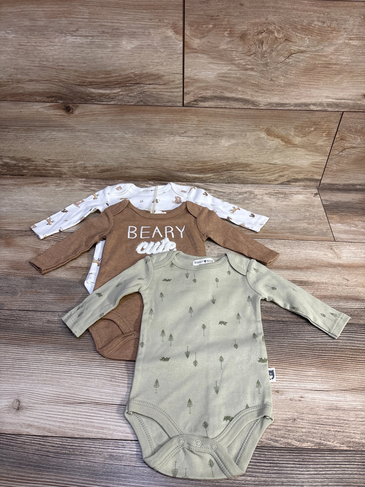 Flat lay of three Rabbit and Bear long-sleeve bodysuits on wood: sage green tree bear print, brown Beary, white rabbit print
