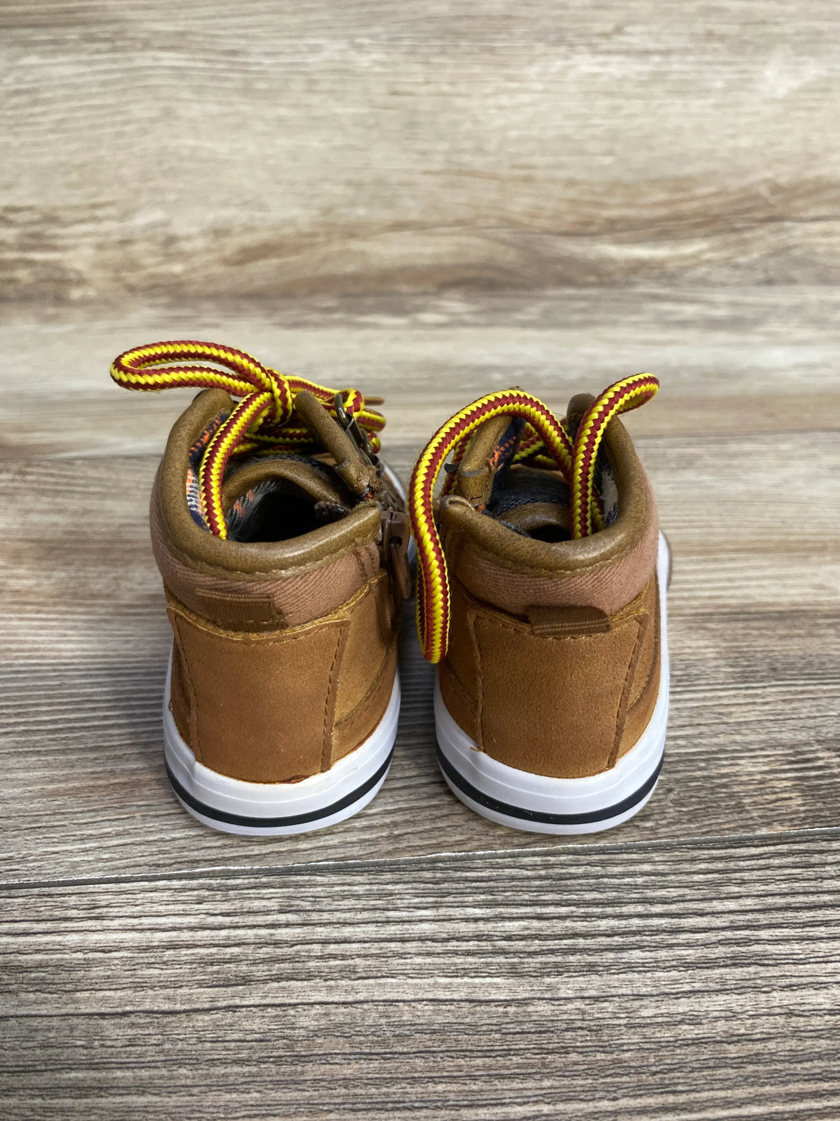 Brown toddler boots with yellow and red laces on wood floor, rear view.
