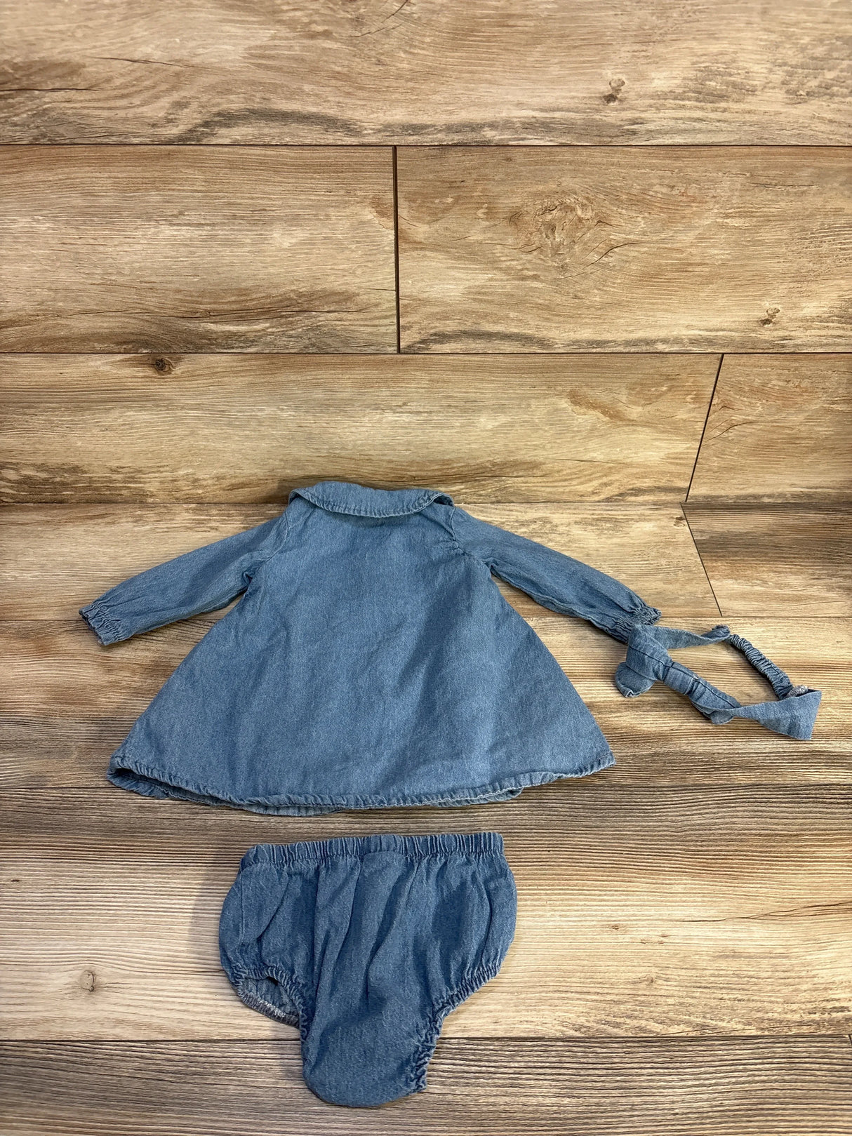 Top-down view of blue 3-piece baby denim set, collared long-sleeve dress, elastic bloomers, knotted headband on wood floor