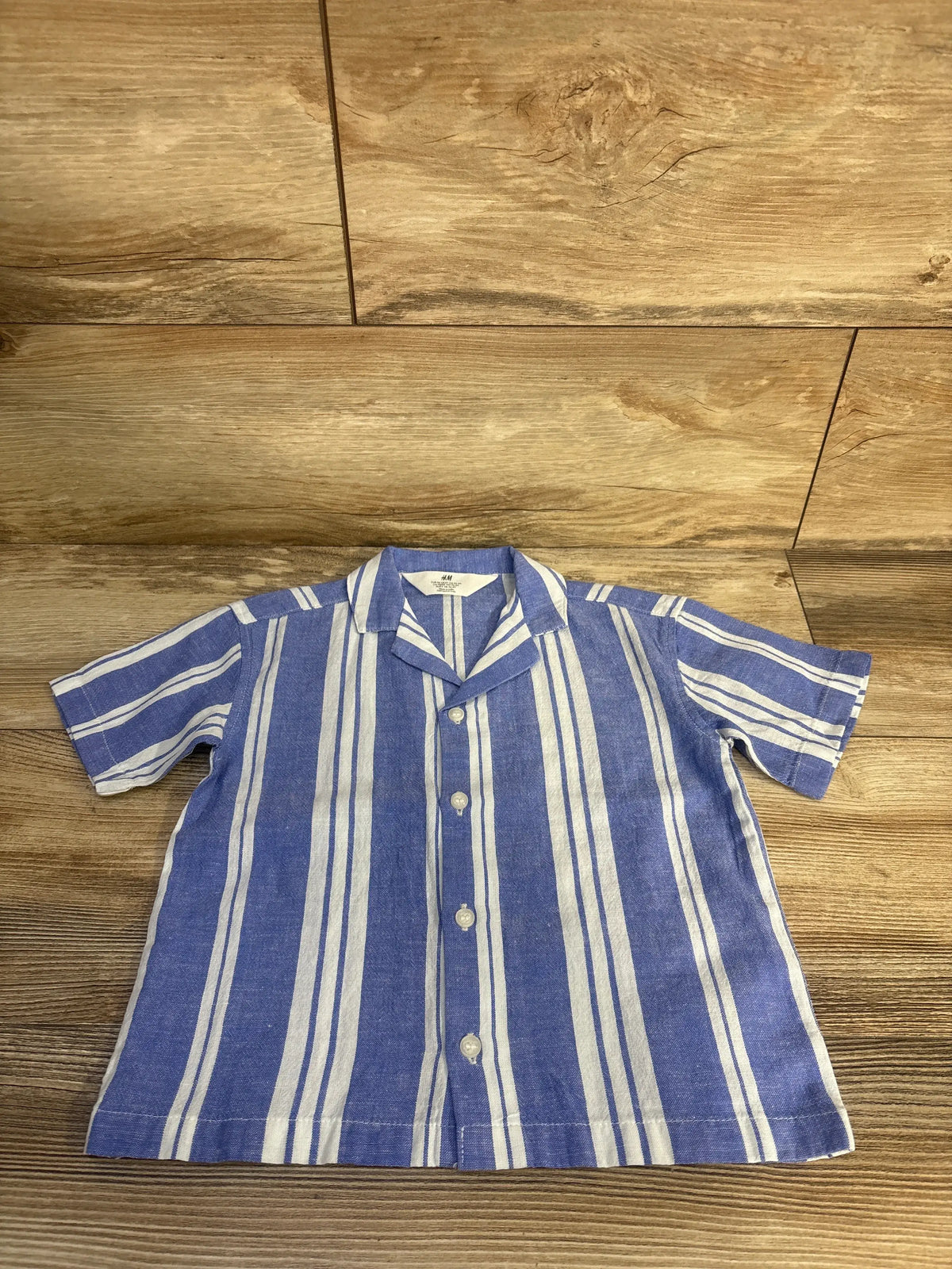 Front view of H&M blue striped button-up resort shirt showcasing short sleeves, resort collar, and buttons on wood-grain background