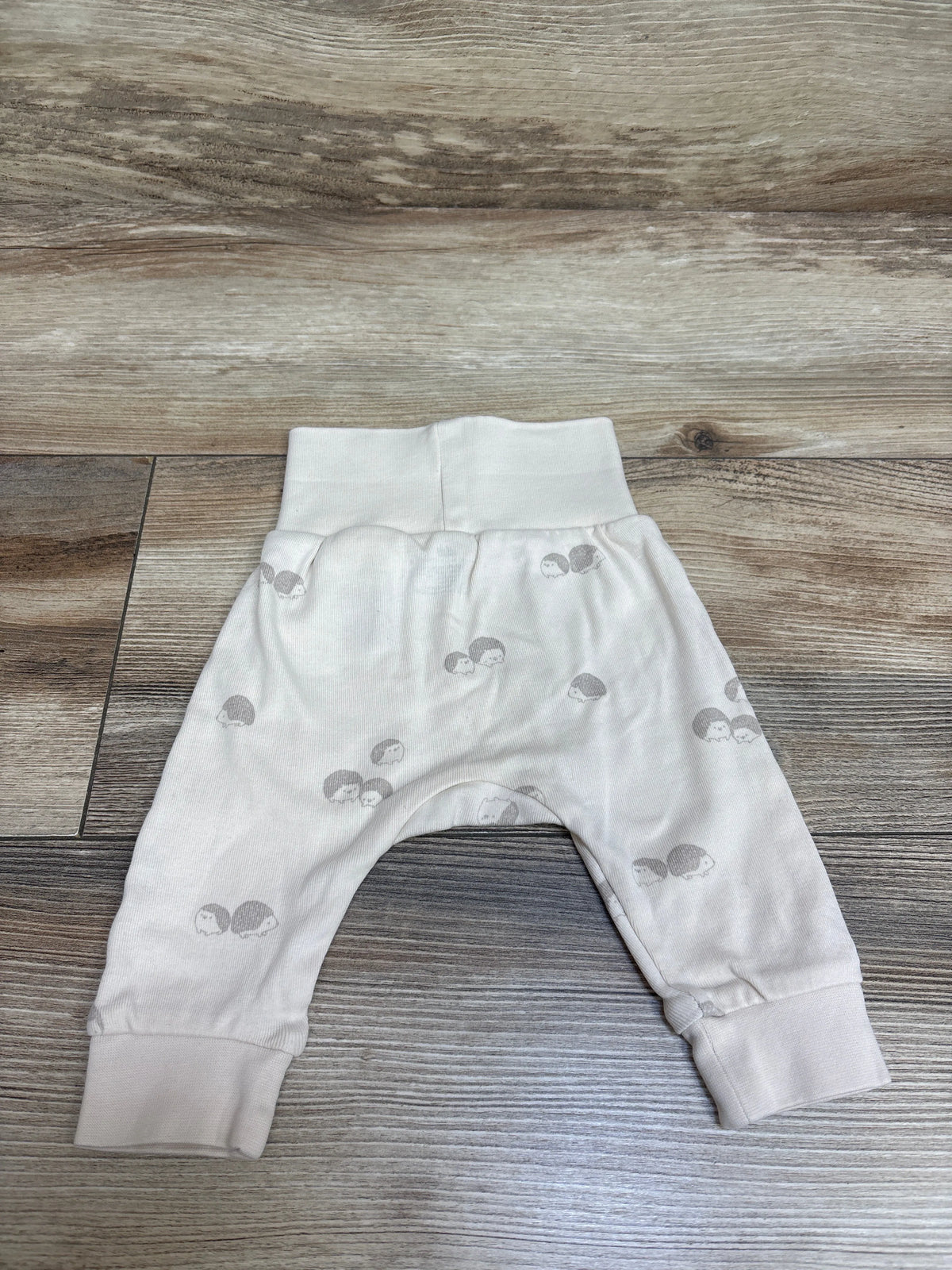 Top-down view of H&M cream foldover-waist pants in newborn size showcasing gray hedgehog pattern and ribbed cuffs on a wood surface