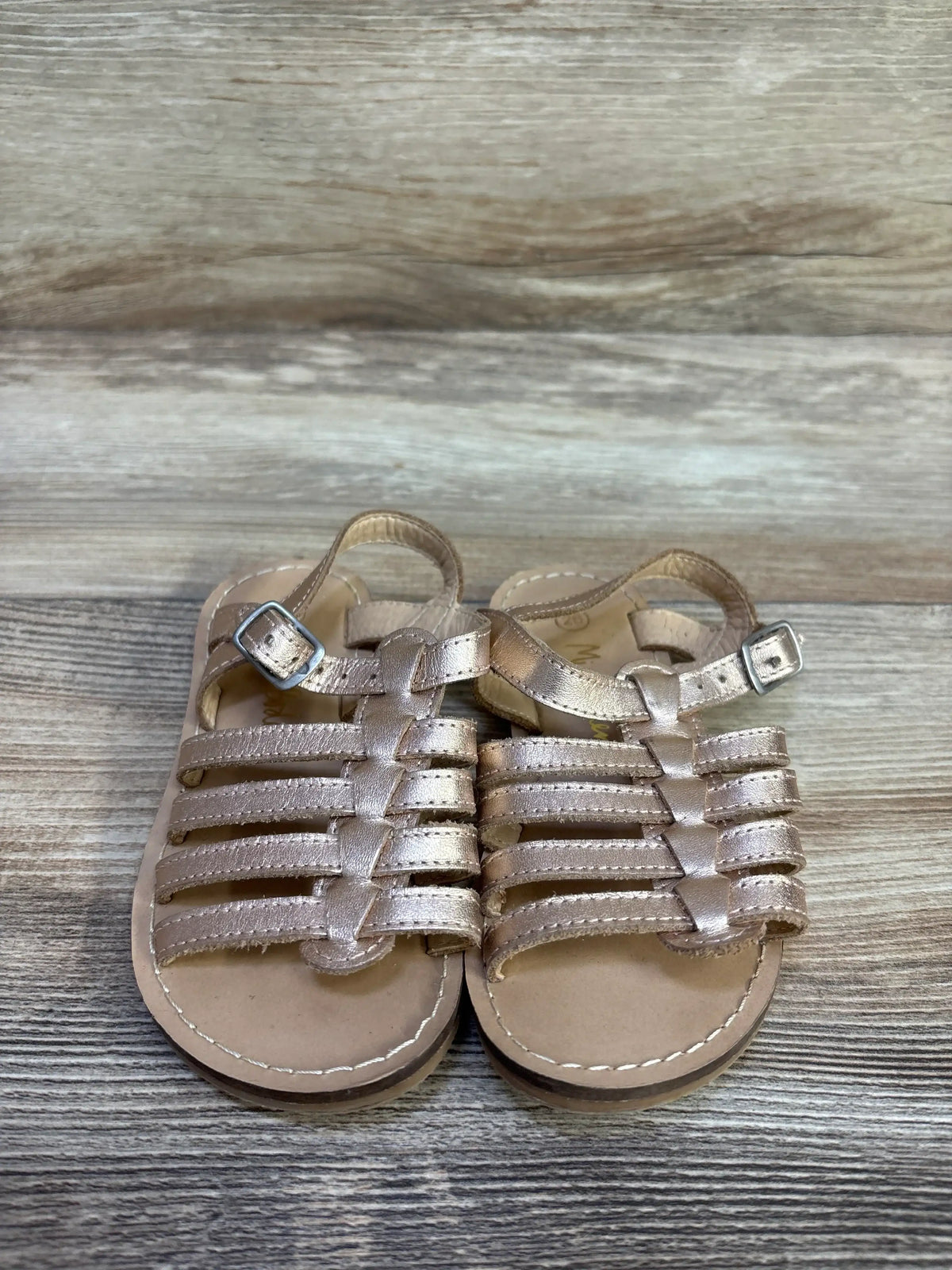 Top view of Mini Boden toddler gladiator sandals in gold showing straps and ankle buckles on wood background