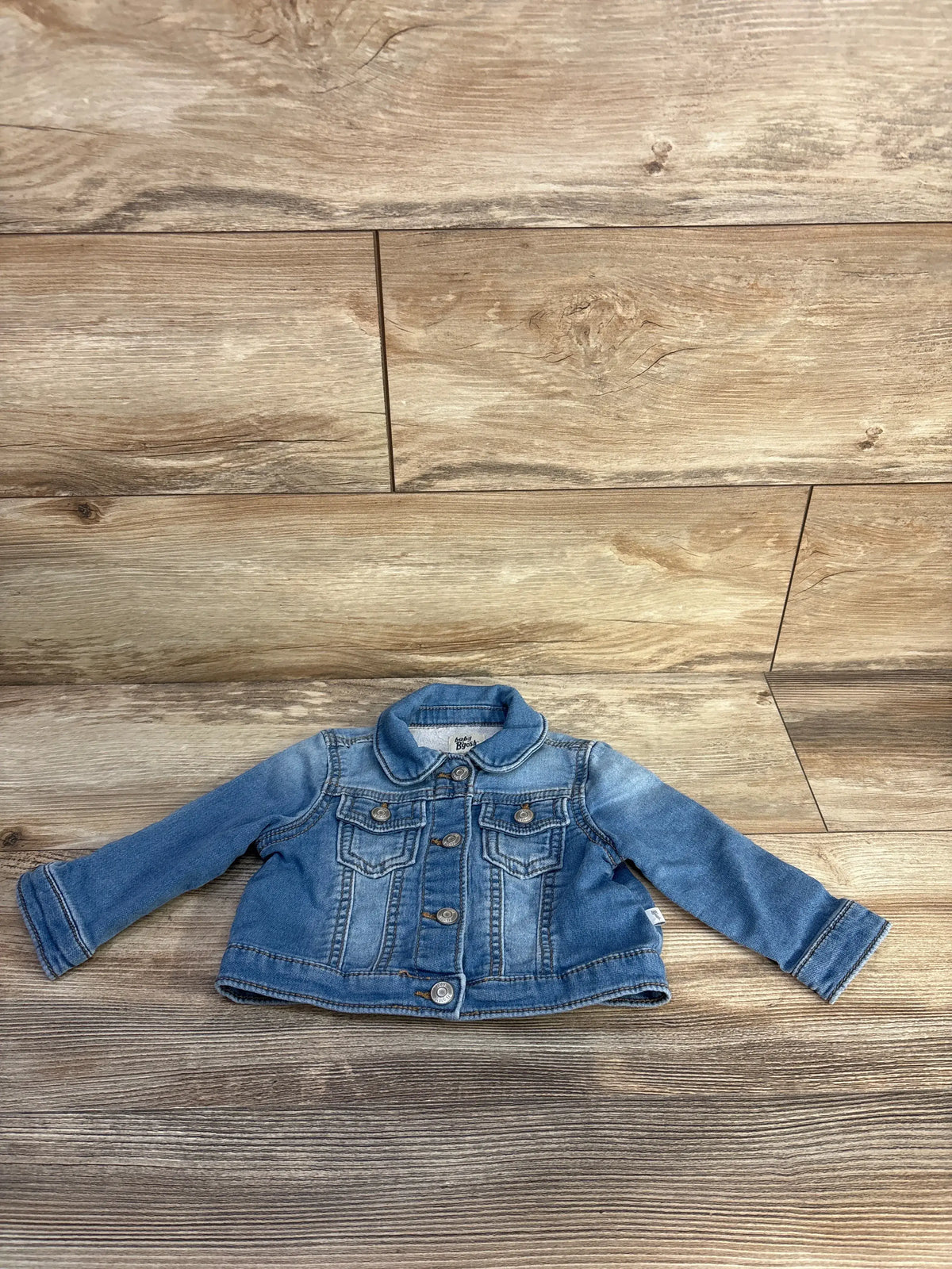 Front view of OshKosh blue denim jacket, size 12 months, showcasing button front and chest pockets with wood floor background
