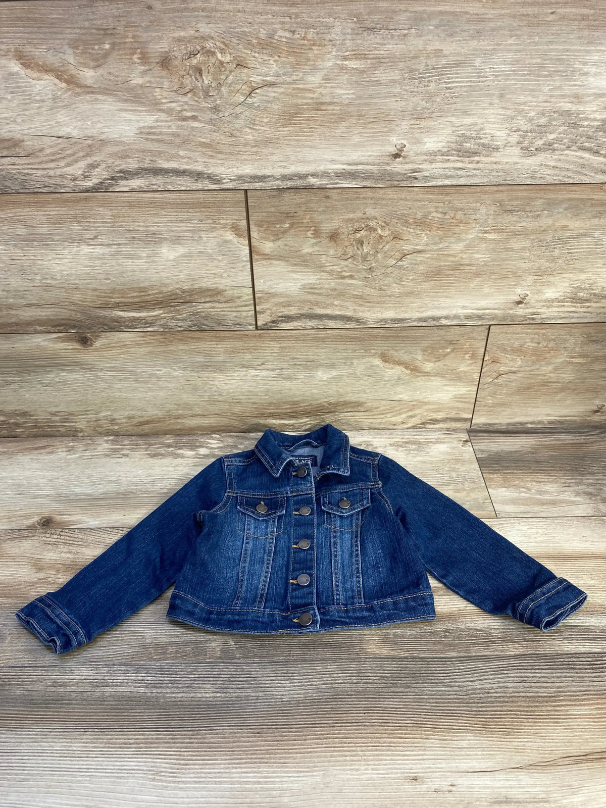 Front flat lay of Children's Place toddler denim jacket size 2T showing button-front closure and chest flap pockets on wood background