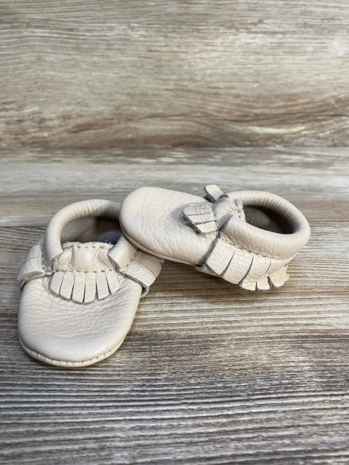 Beige leather baby moccasins with fringe on a wooden background