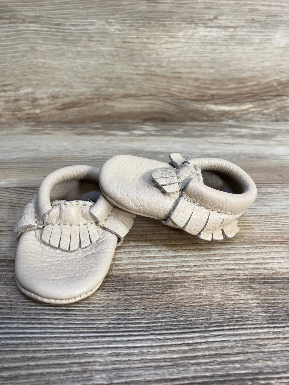 Beige leather baby moccasins with fringe on a wooden background