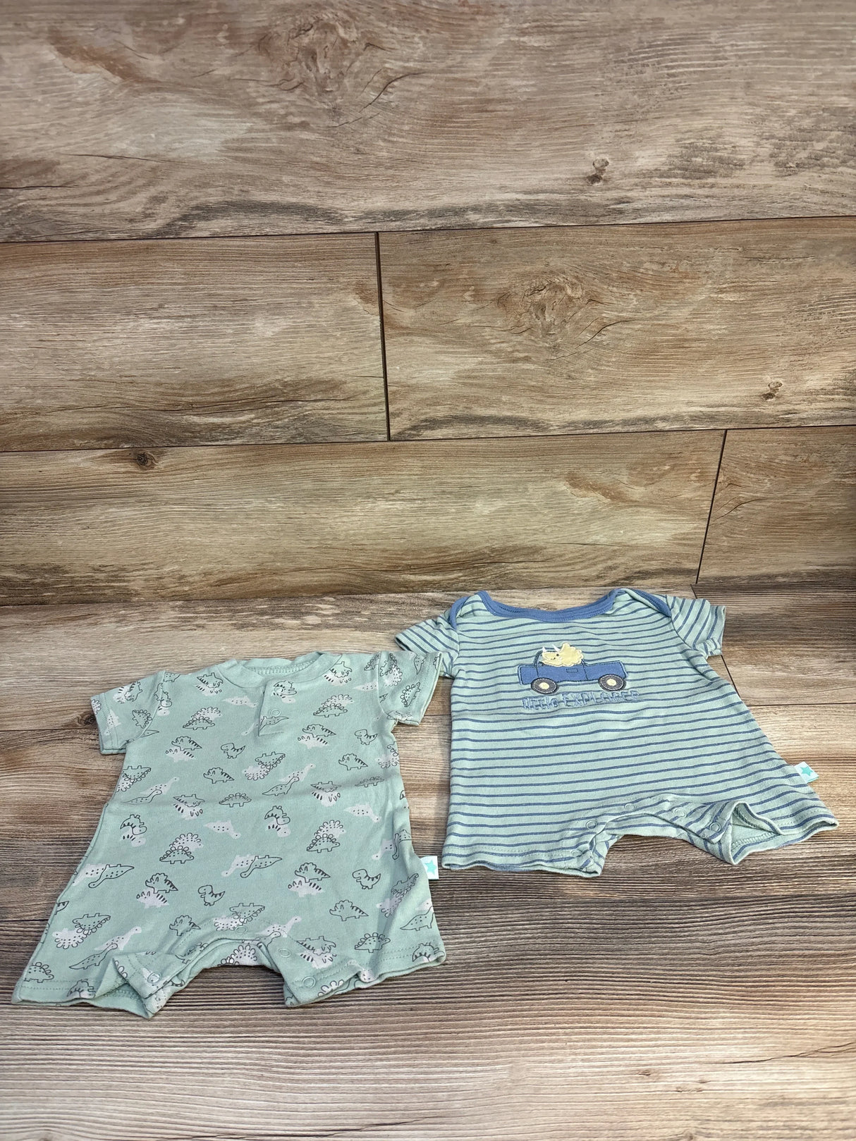Two baby boy rompers, one with dinosaur print and one blue striped with truck, on wood background.