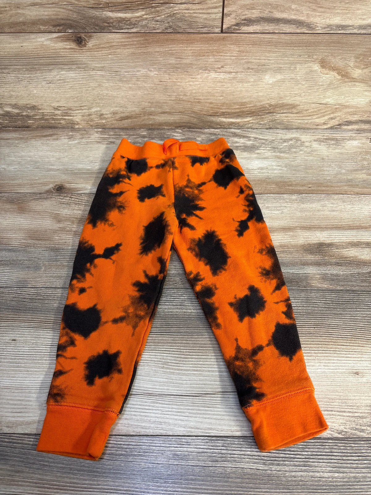 Front view of orange Halloween joggers with black tie-dye pattern, elastic waistband, and cuffed ankles on a wood background