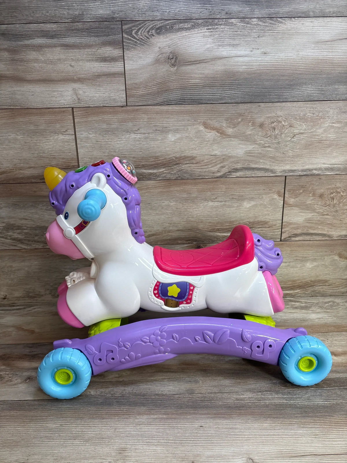 Side view of VTech Prance And Rock Learning Unicorn with pink seat, interactive buttons and blue wheels on wood floor
