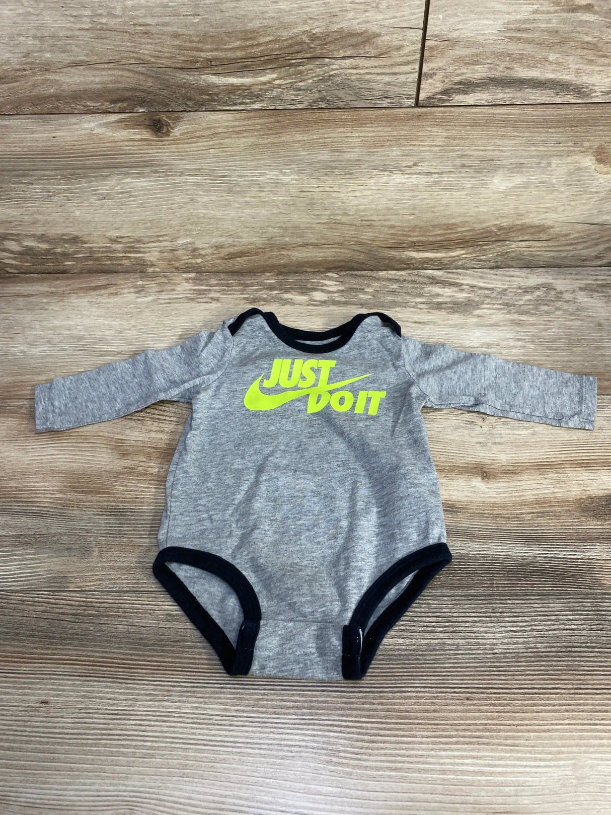 Flat lay of grey Nike long-sleeve baby bodysuit (3–6M) showing neon yellow JUST DO IT logo, black trim, and snap crotch on wood floor