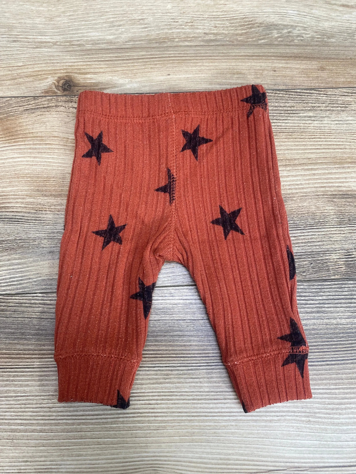 Front view of Grayson Collective ribbed newborn pants in rust showing black stars, elastic waist and cuffs on wood surface